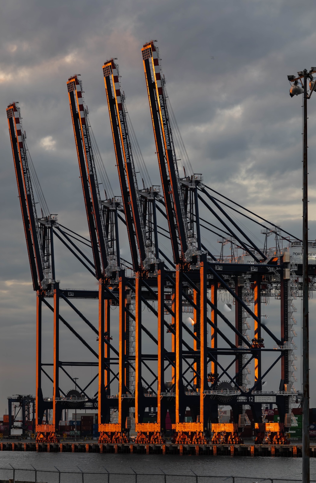 Four container-terminal gantry cranes at Port Liberty Terminals lit orange by the setting sun against a cloudy sky.