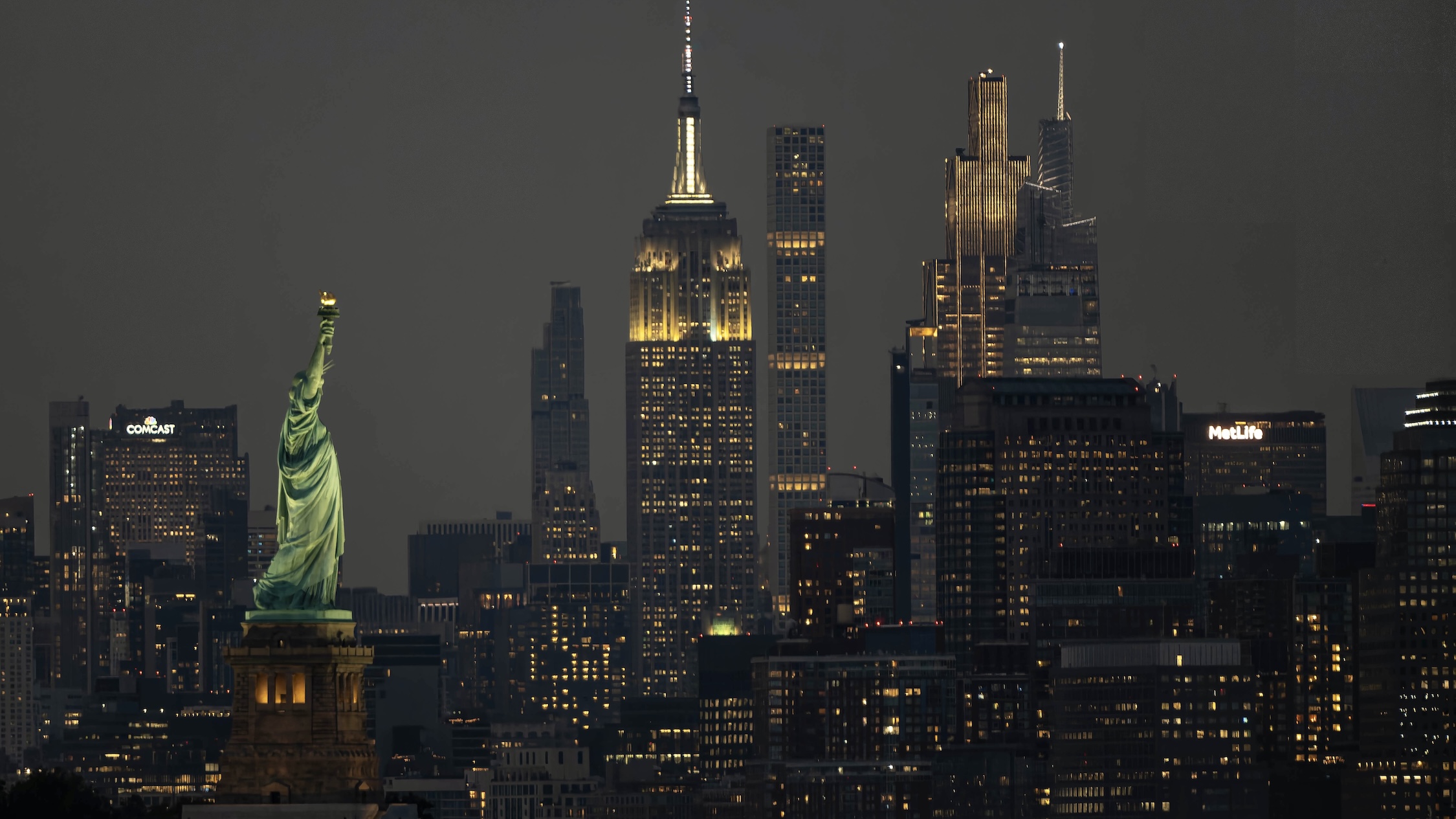 The Statue of Liberty in the foreground with the illuminated Empire State Building and Midtown Manhattan skyscrapers behind it at night.