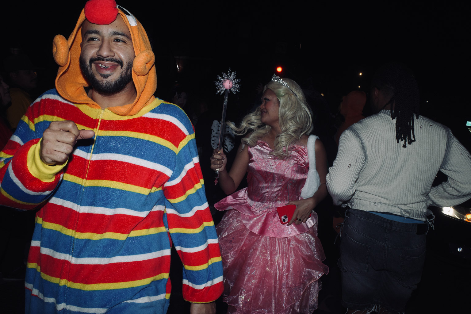 A man in a colourful striped onesie smiles and points while a woman in a pink princess dress stands beside him, holding a wand. The scene is set at night with dim lighting.