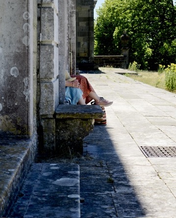 Two women sits on a stone bench in dappled sunlight against a historic building's wall.