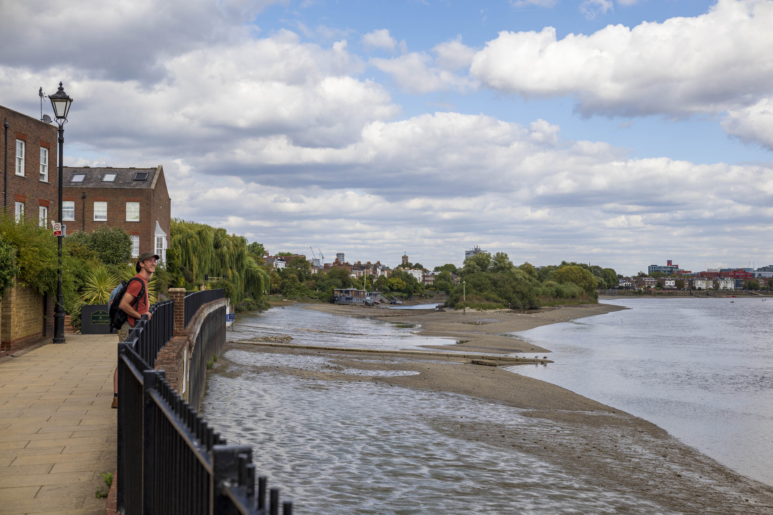 A man leans on a riverside railing overlooking the Thames at low tide, with brick townhouses and a cloudy sky.