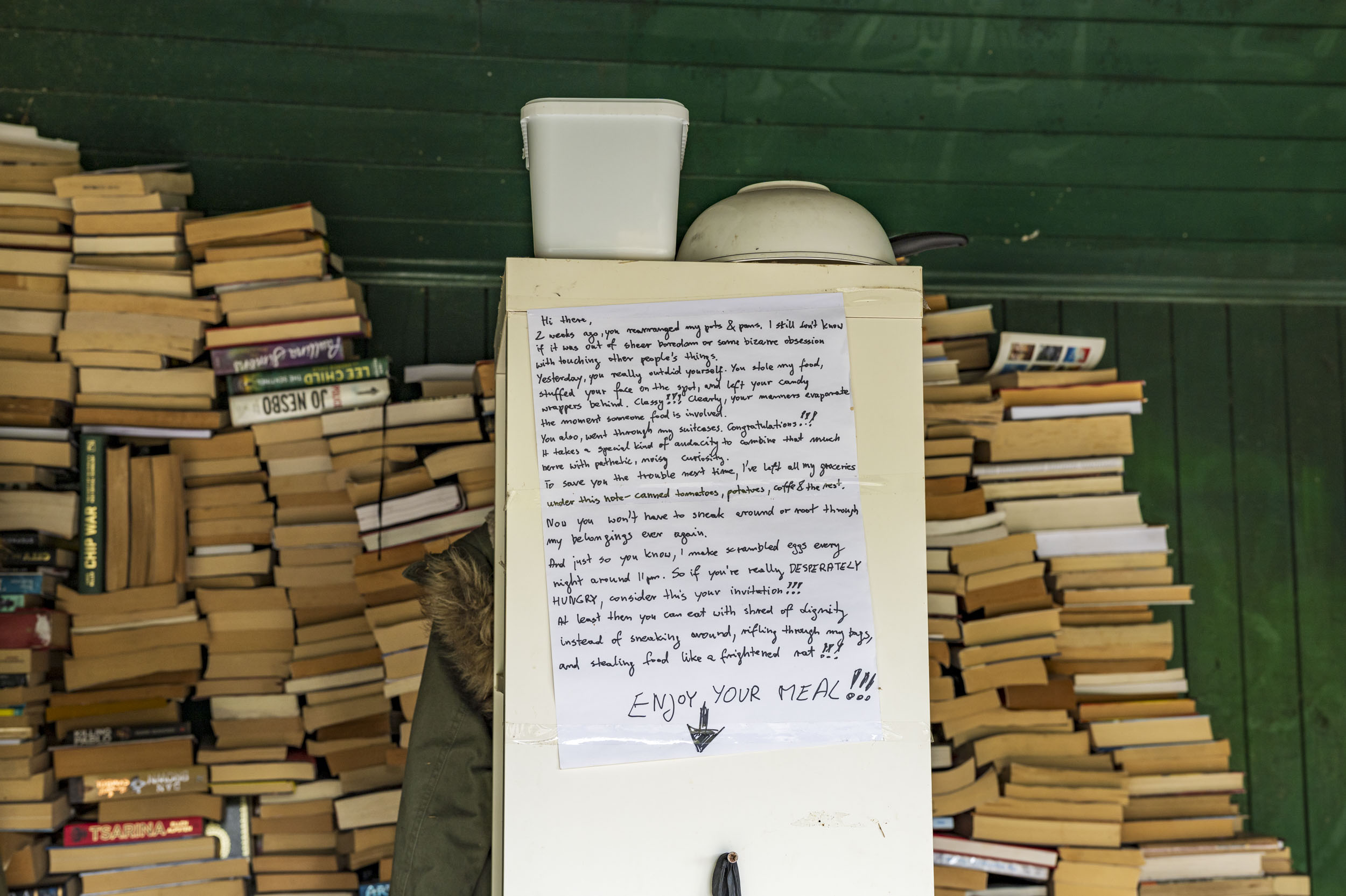 A handwritten note taped to a cupboard door, surrounded by stacks of secondhand books in a green-painted shed.