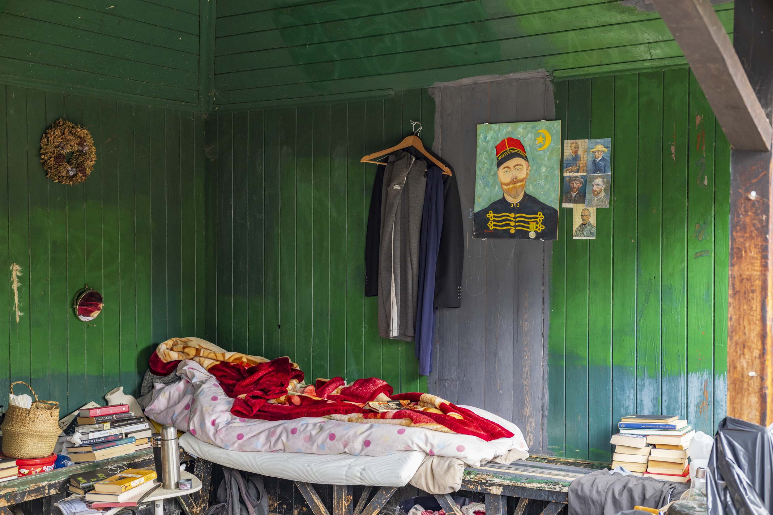 An eccentric shed interior with a red blanket on a bed, coats hanging, and a portrait of a Zouave soldier on the wall.