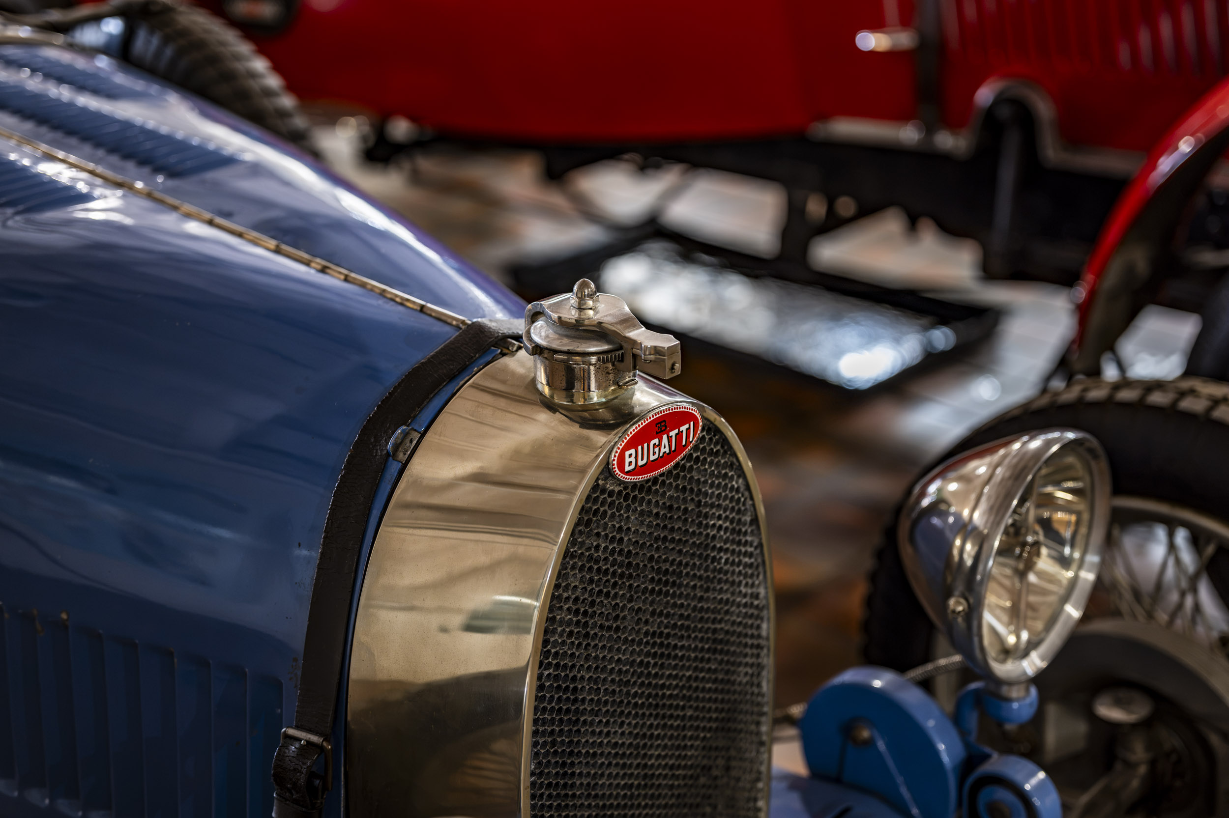 The badge and honeycomb radiator grille of a vintage blue Bugatti racing car, with other classic cars blurred behind.