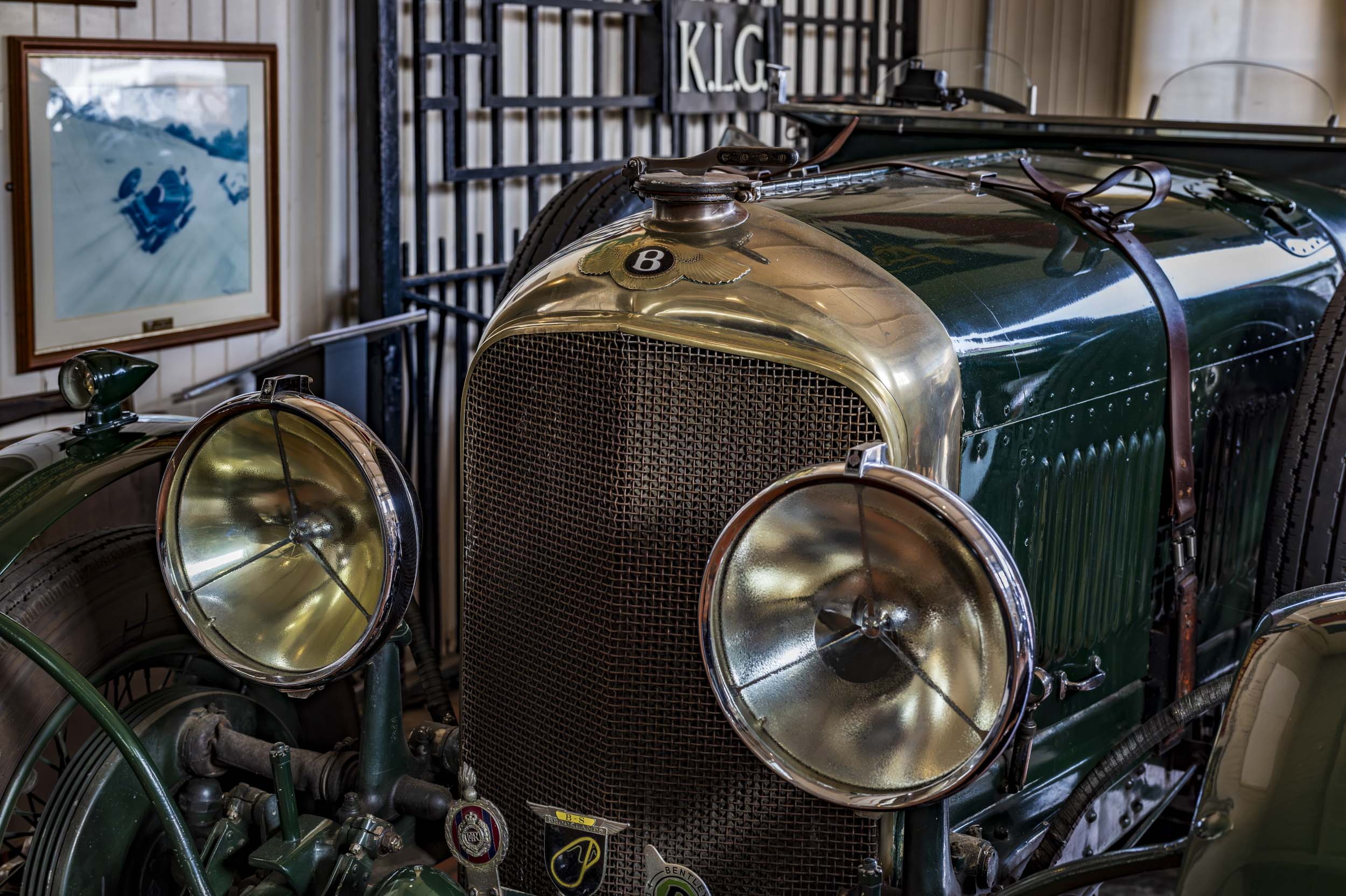 The brass radiator grille and headlamps of a vintage green Bentley, with a framed racing photograph in the museum background.