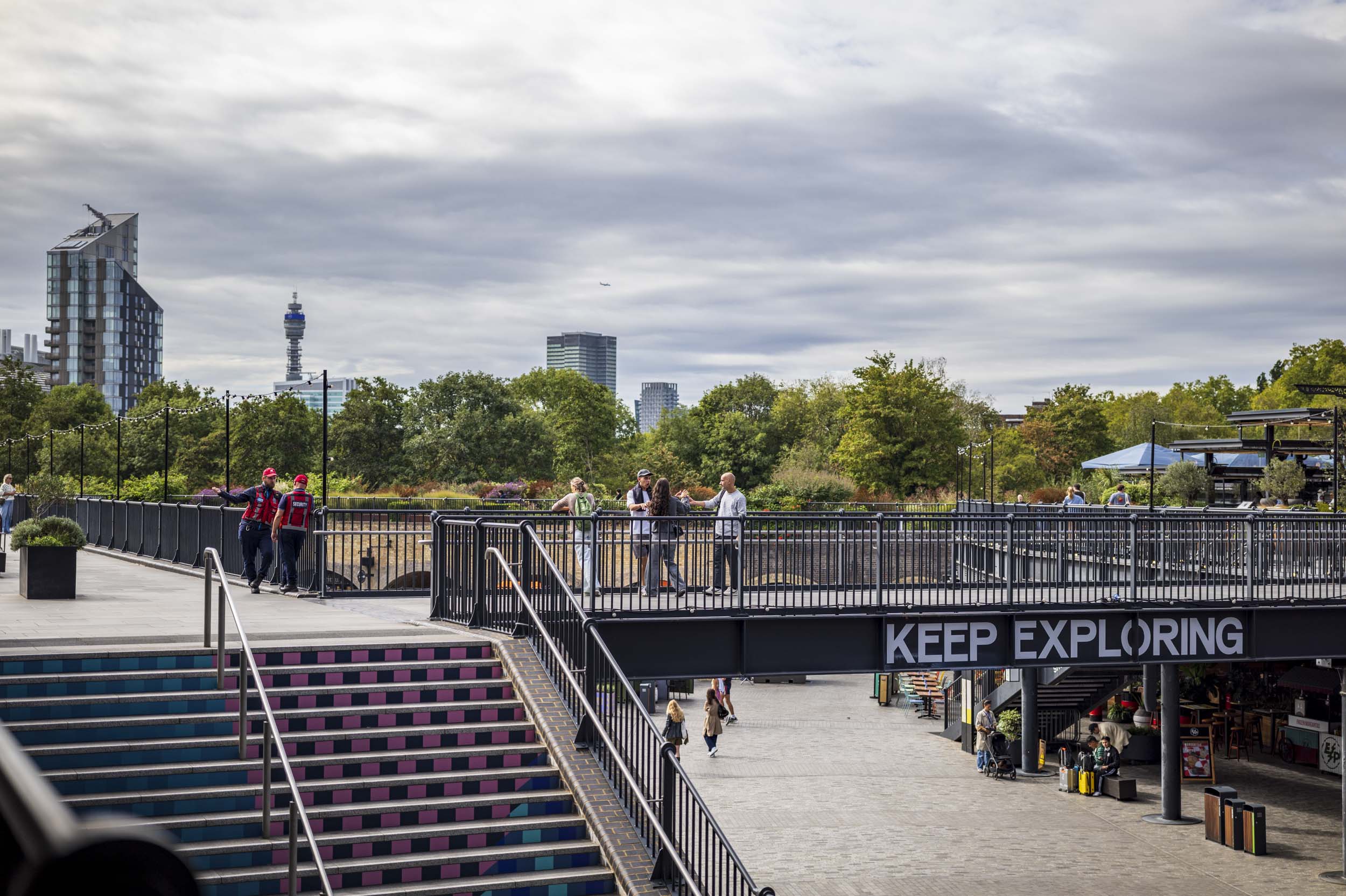 An elevated pedestrian bridge marked ‘KEEP EXPLORING’ over disused railway tracks, with people chatting and London’s BT Tower visible on the skyline behind.
