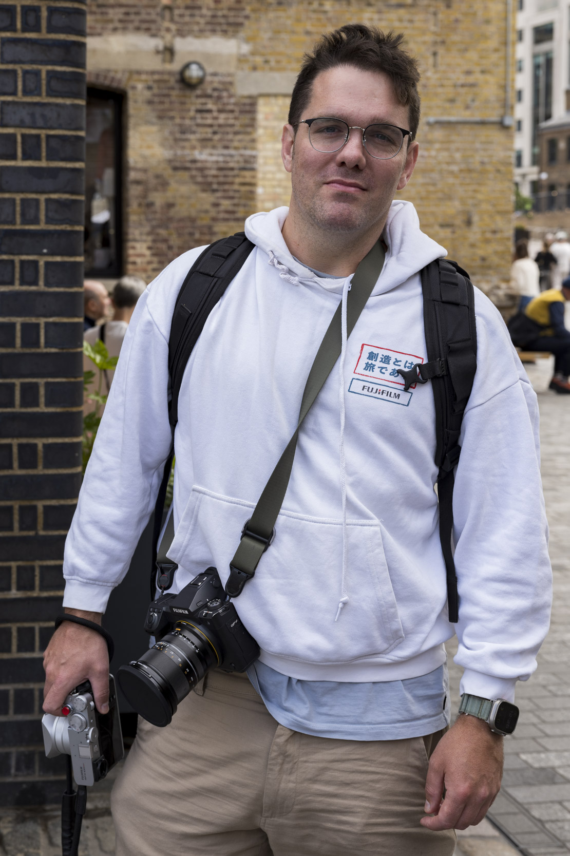 A man wearing a Fujifilm-branded hoodie holds a camera on a city street, brick building behind him.