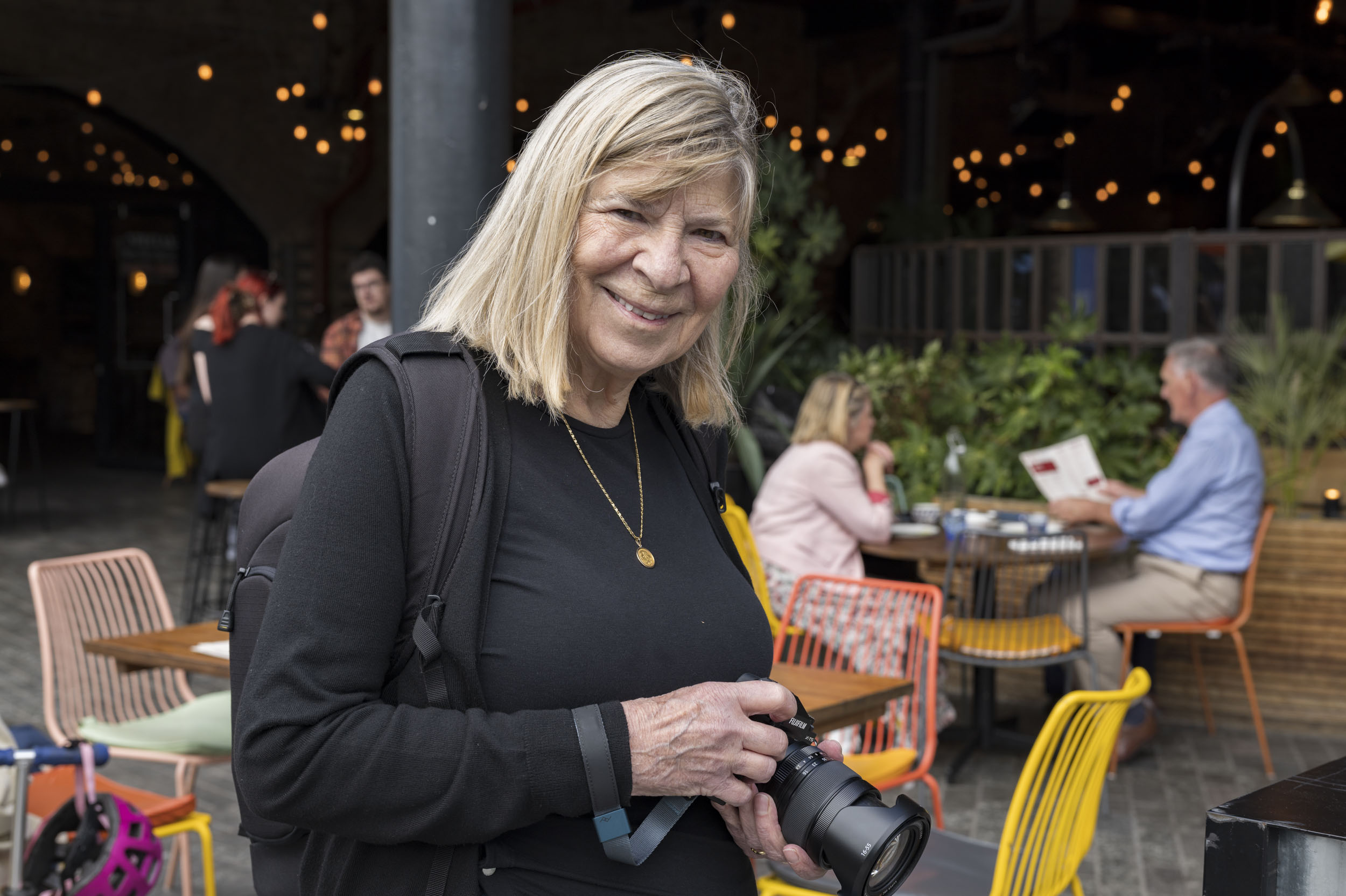 A smiling woman holds a Fujifilm camera in a café courtyard strung with fairy lights.