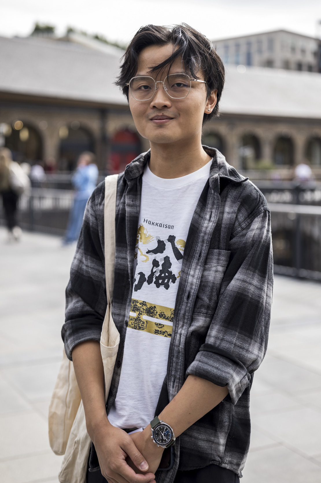 A young man wearing glasses and a plaid shirt over a graphic T-shirt, standing outdoors in a covered market.