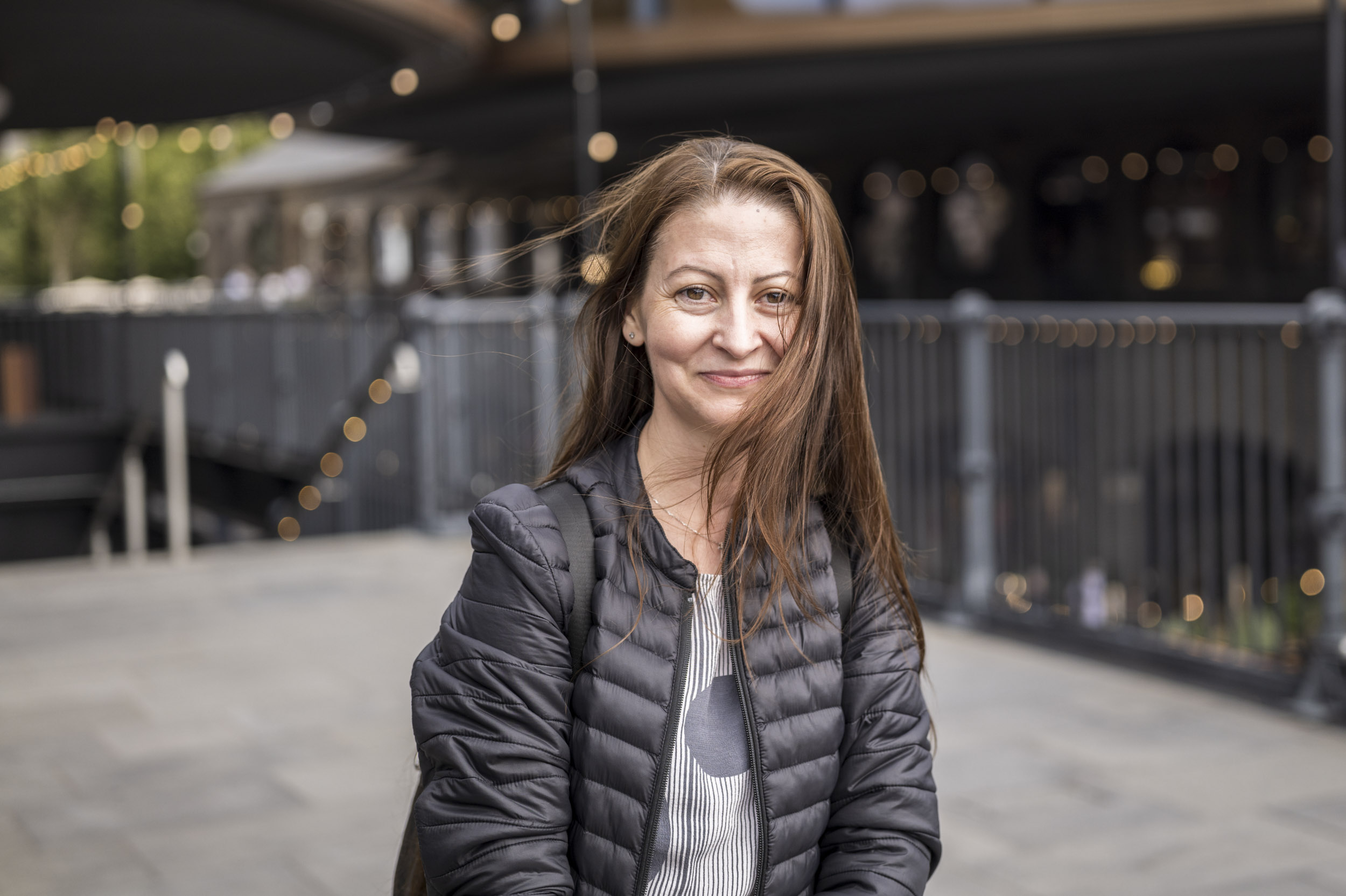 A woman smiles for a portrait with her hair blowing in the wind, a canalside market blurred behind her.