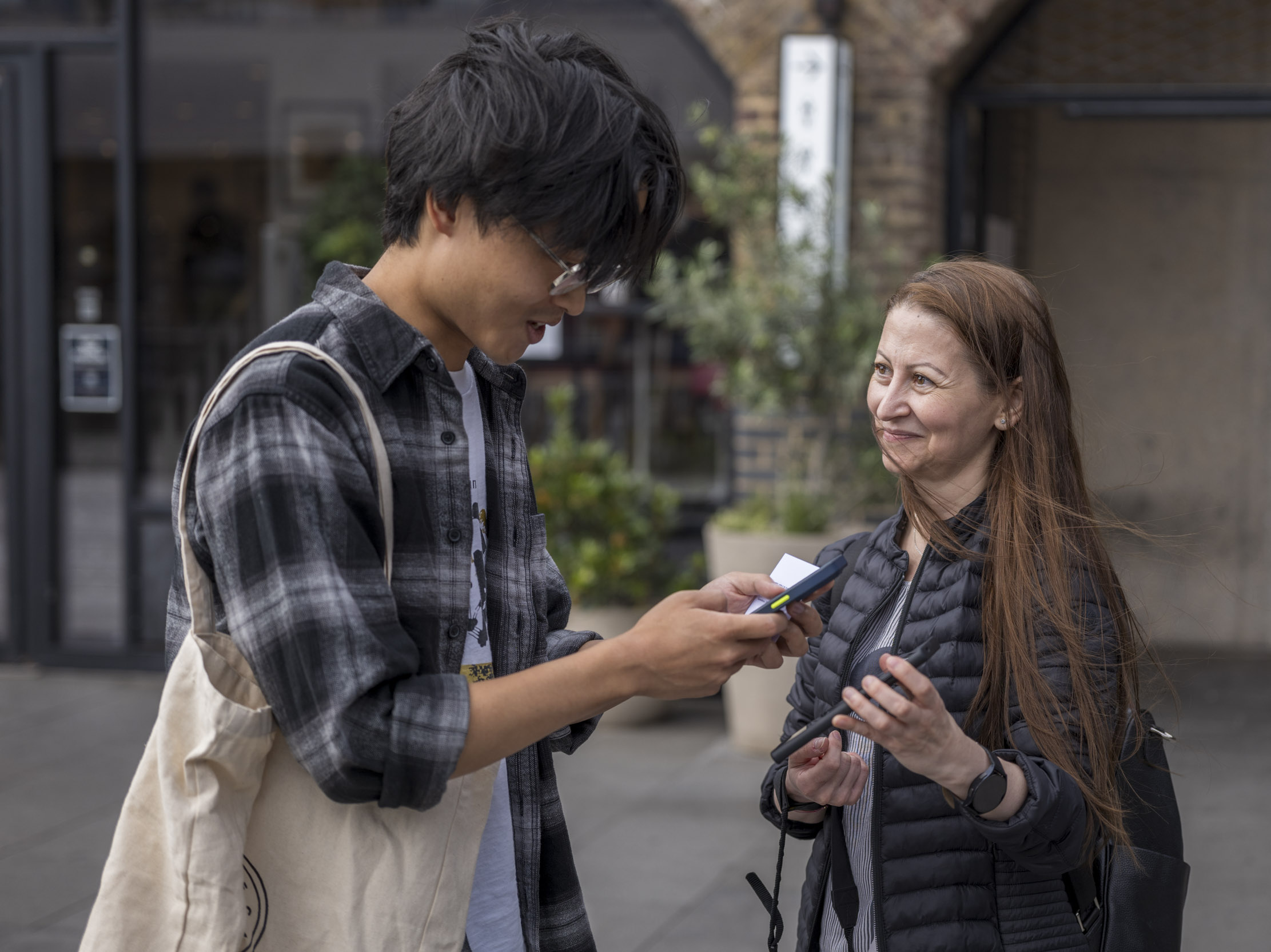 Two people chat on a street, one showing something on a phone, with shopfronts blurred behind them.