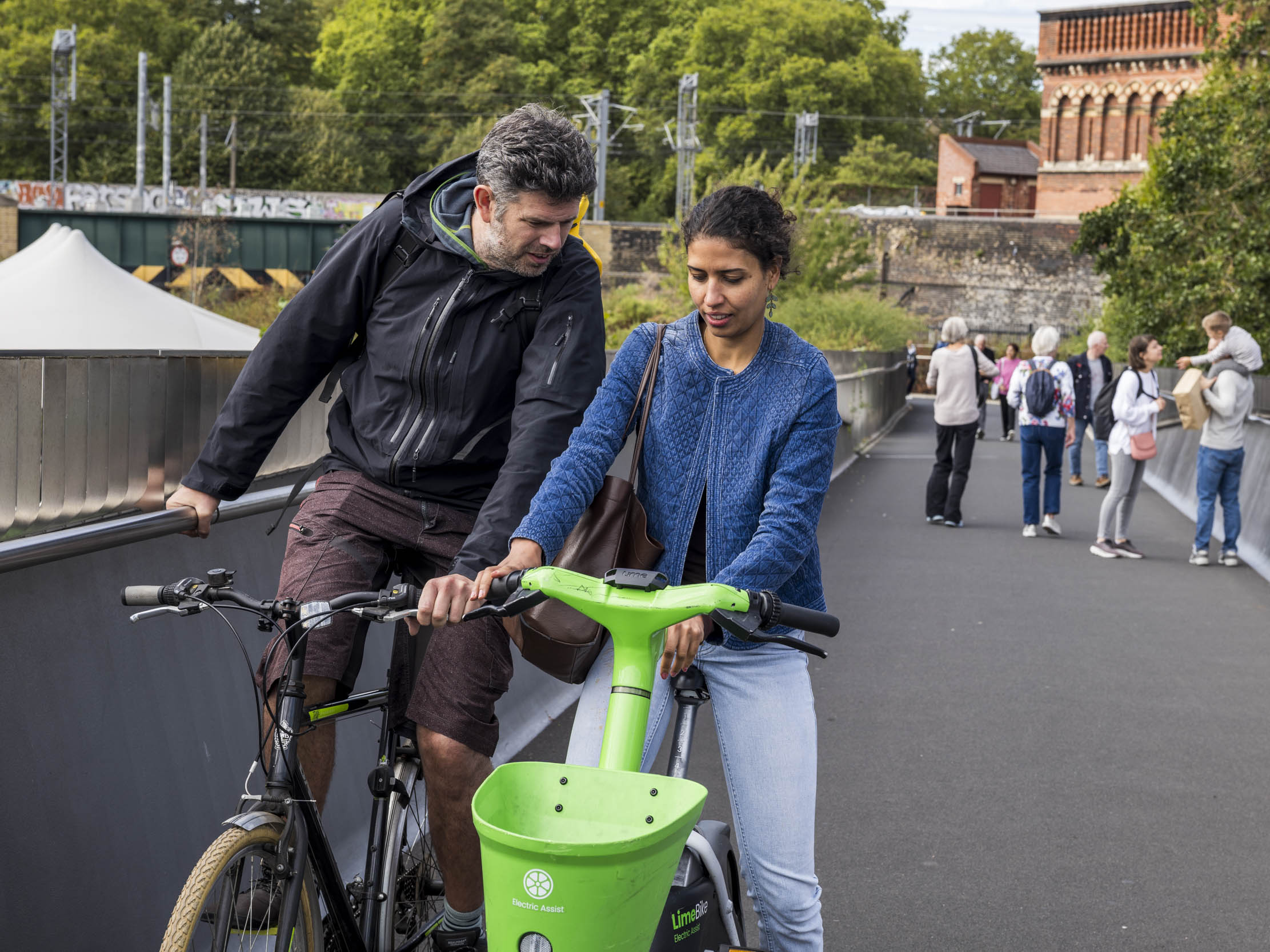 A cyclist and a woman with a Lime e-bike pause to chat on a pedestrian bridge above a railway line.