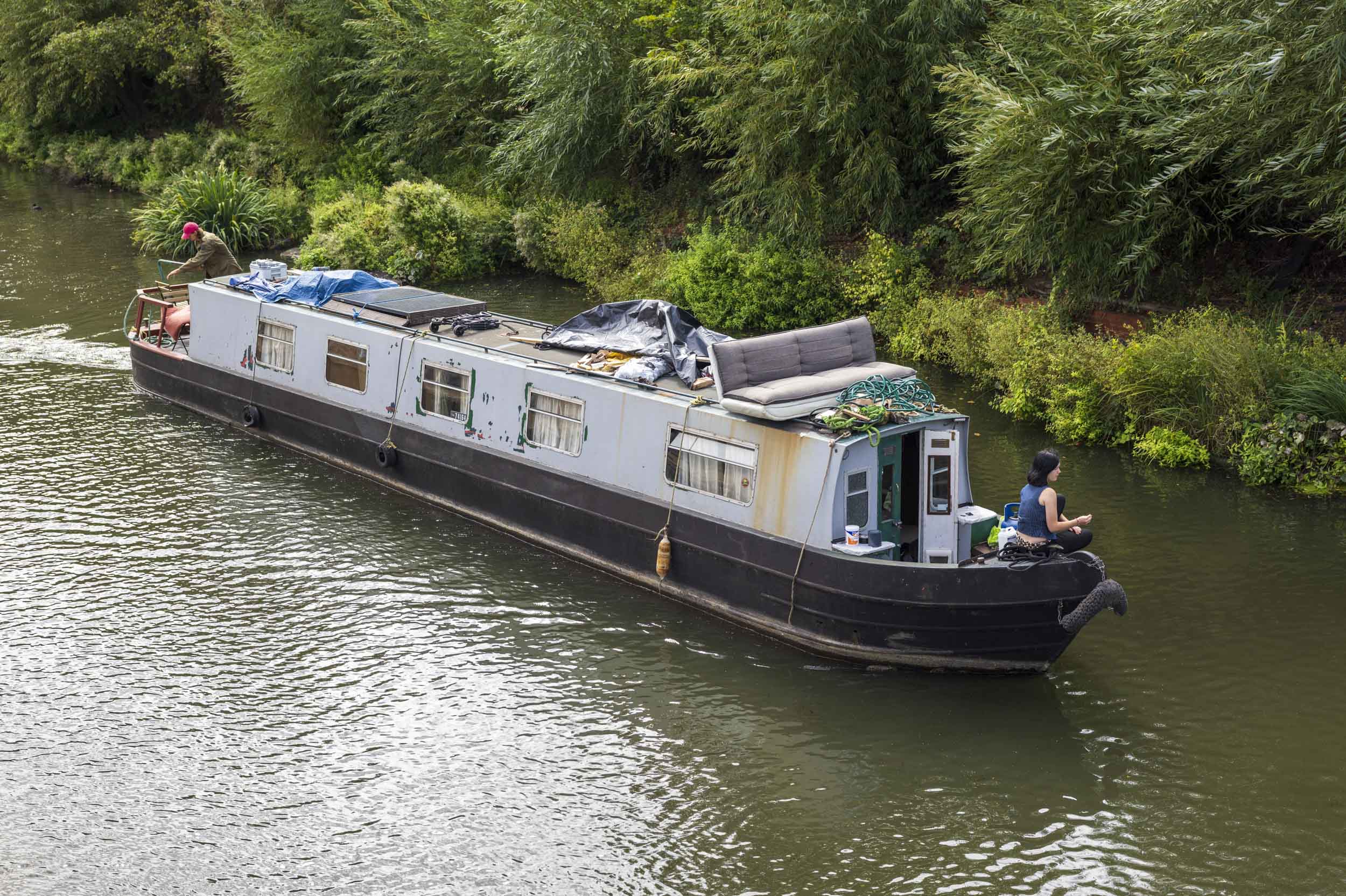 A well-lived-in narrowboat, its roof piled with belongings, cruises a tree-lined canal with a woman seated at the stern.