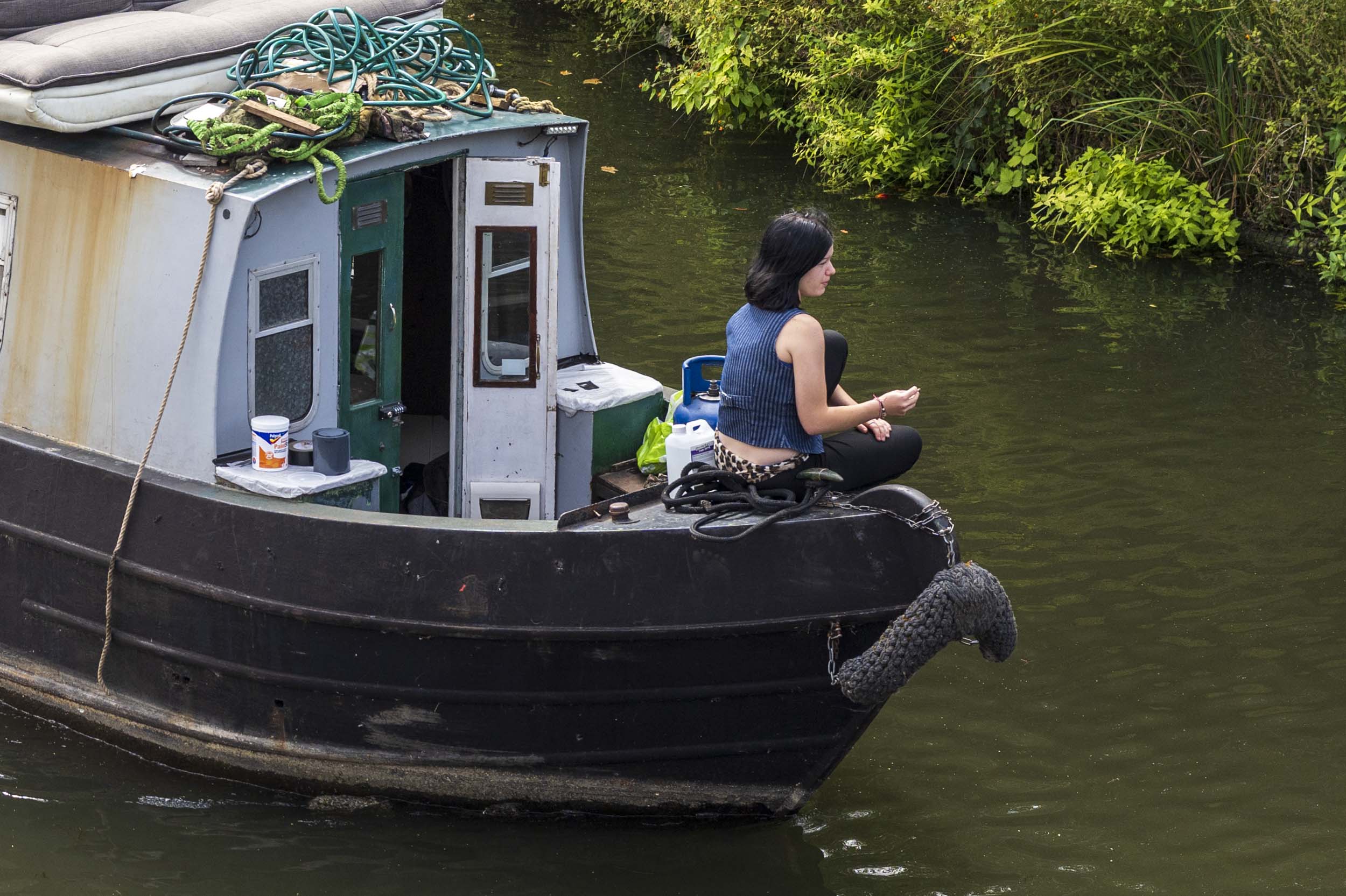 A close crop of a woman sitting cross-legged on the bow of a narrowboat, looking out over the water.