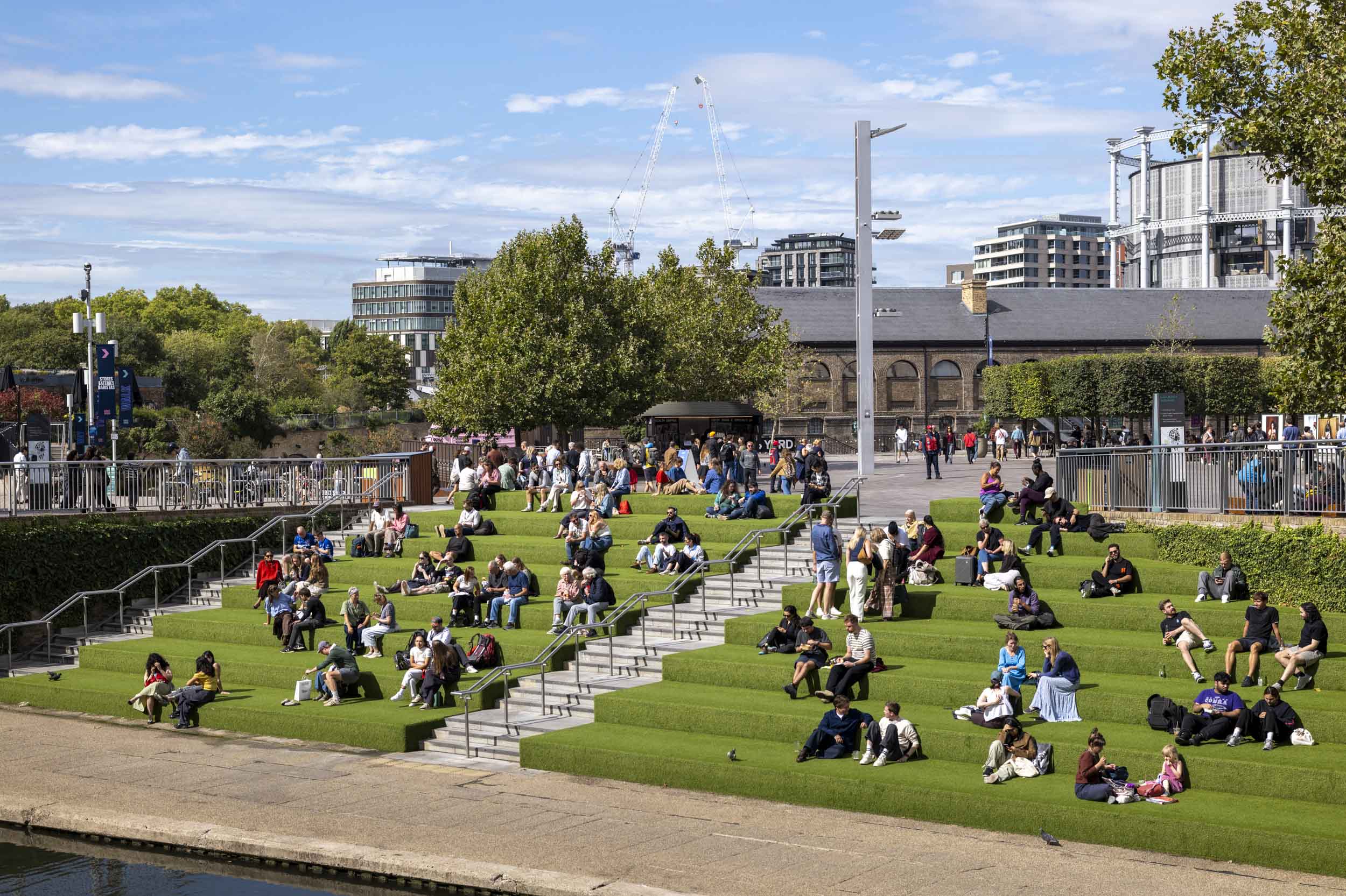 Crowds relax on the grass-covered amphitheatre steps of Granary Square at King's Cross, with the canal in front and the gasholder apartments behind.