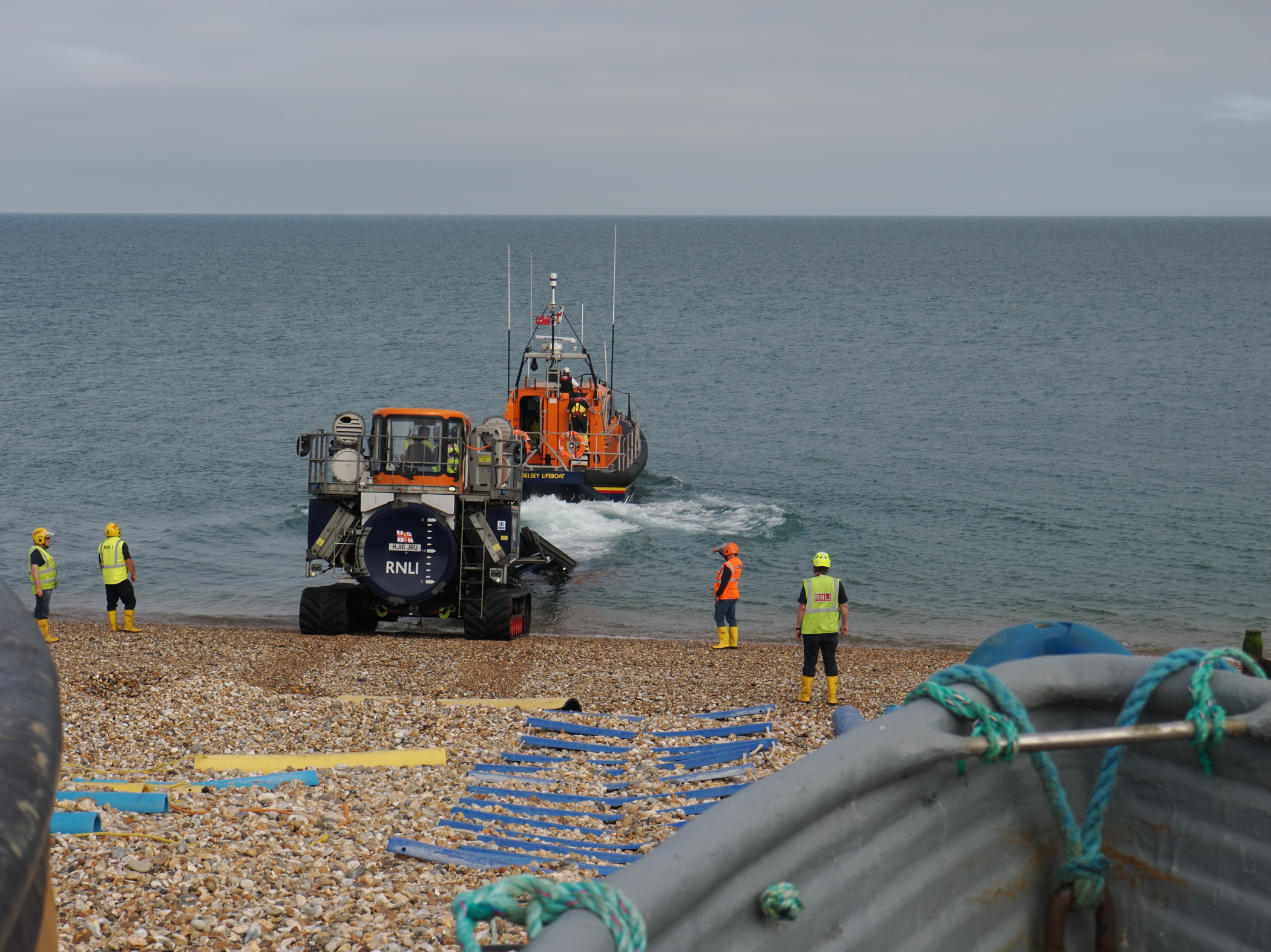 A lifeboat approaching the shore with rescue workers in high visibility jackets on a pebble beach. The scene features a range of safety equipment and a coastal landscape.