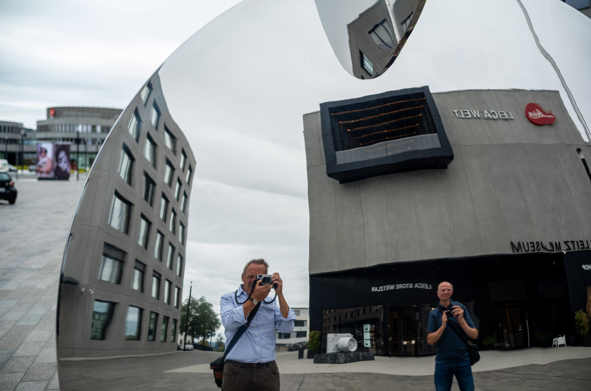 Leica Wetzlar visit: Two men photographing with rangefinders camera, doubled in reflection by a large curved mirror sculpture outside the Leica Welt building.