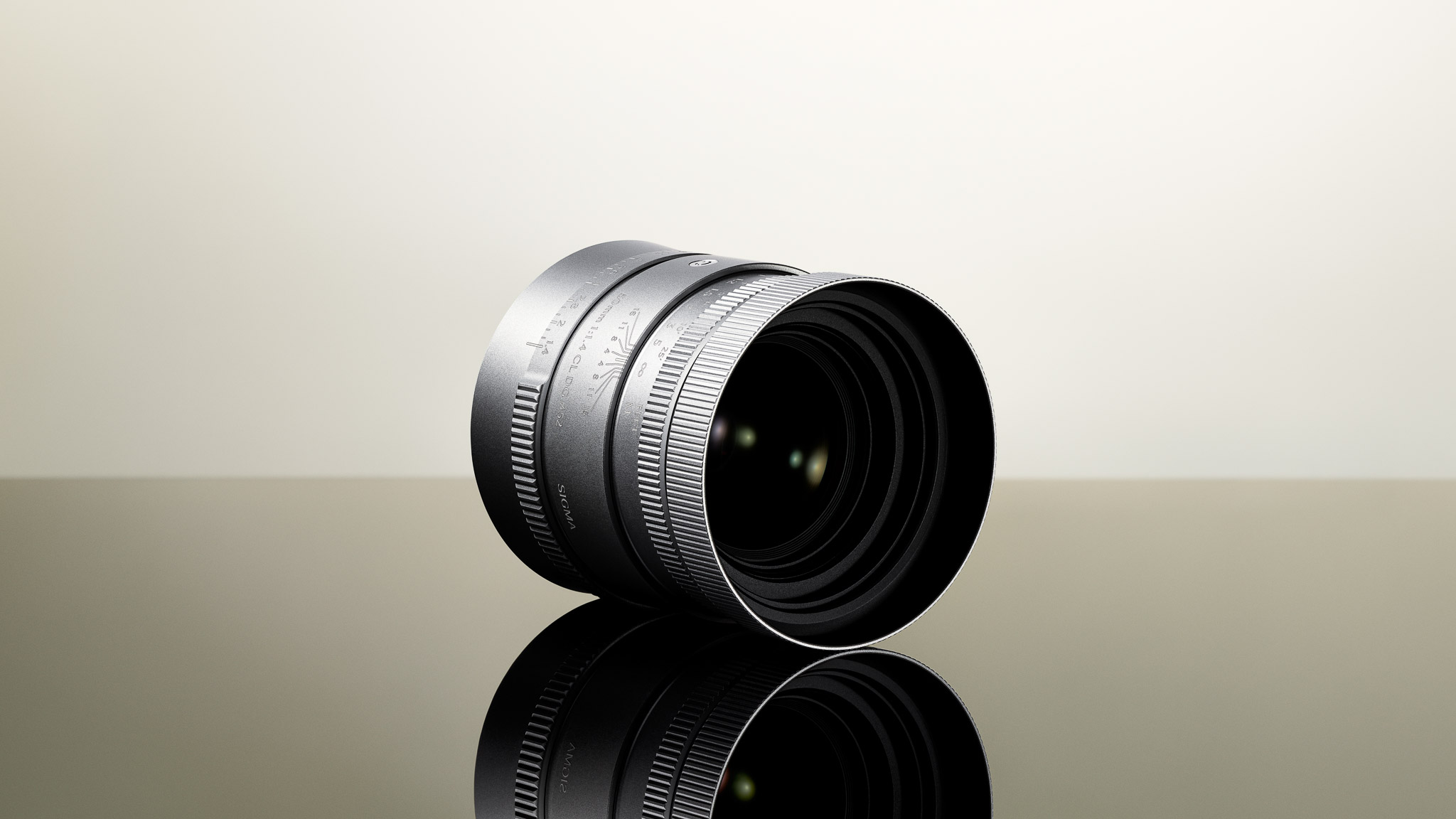 A silver Sigma 50mm f/1.4 CL lens seen at an angle, showing its knurled hood, focus ring and aperture ring.