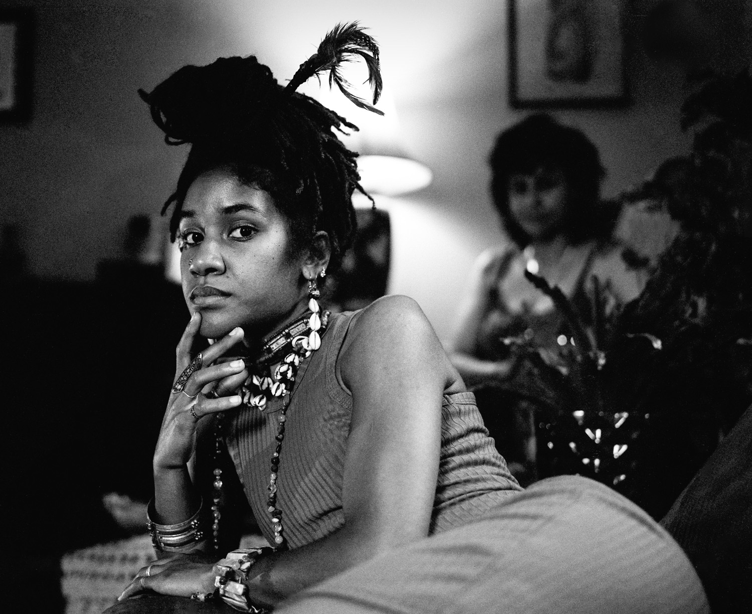 Black-and-white photograph from the Clayton Sisterhood Project: a young woman with locs topped by a feather rests her chin on her hand, while another woman sits blurred in the background beside a lamp.