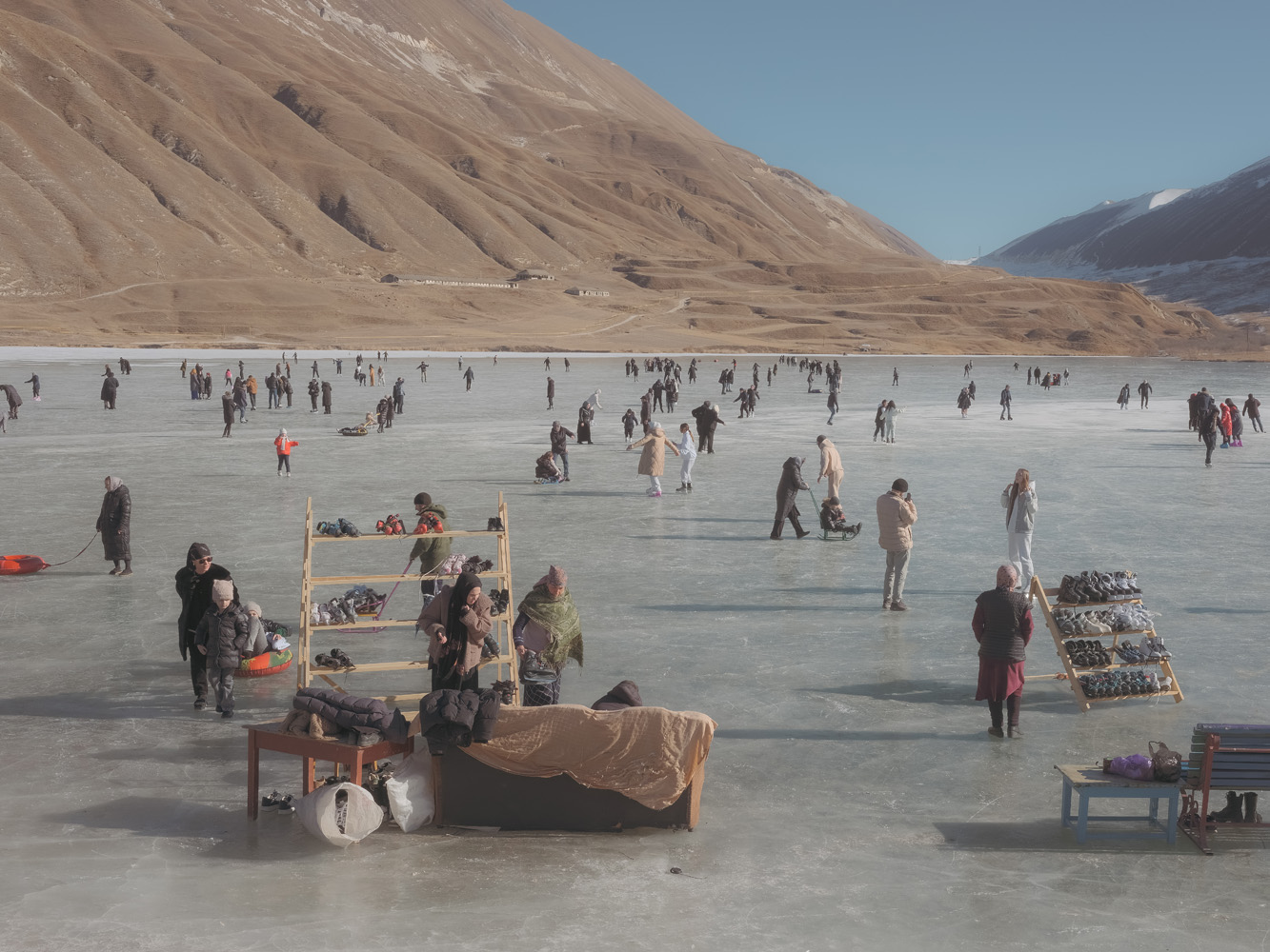 Crowds skating on a frozen mountain lake in Dagestan, with skates for hire on wooden racks in the foreground and bare brown slopes behind.