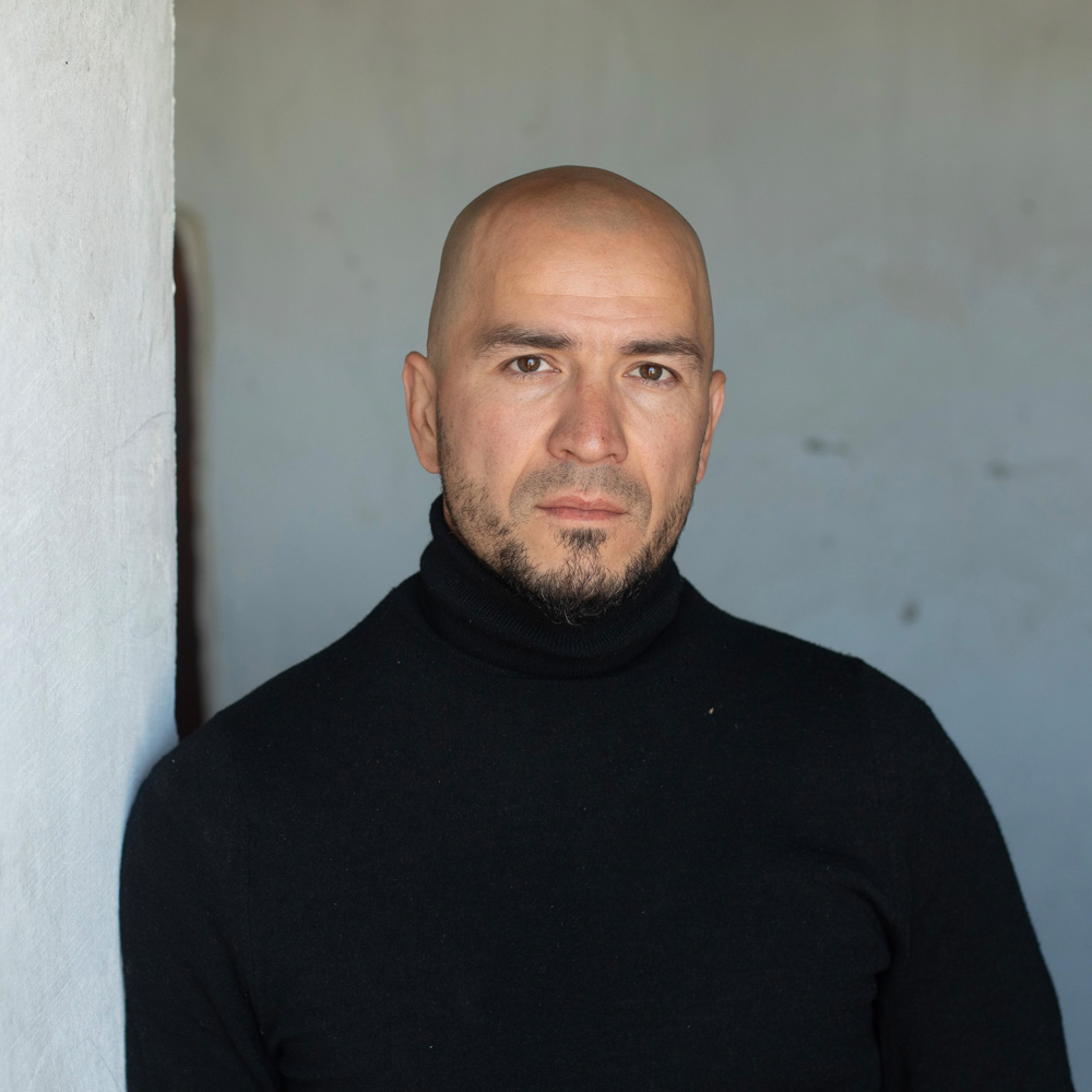 Portrait of Damir Faizulin, bald and bearded, in a black roll-neck jumper beside a white wall.