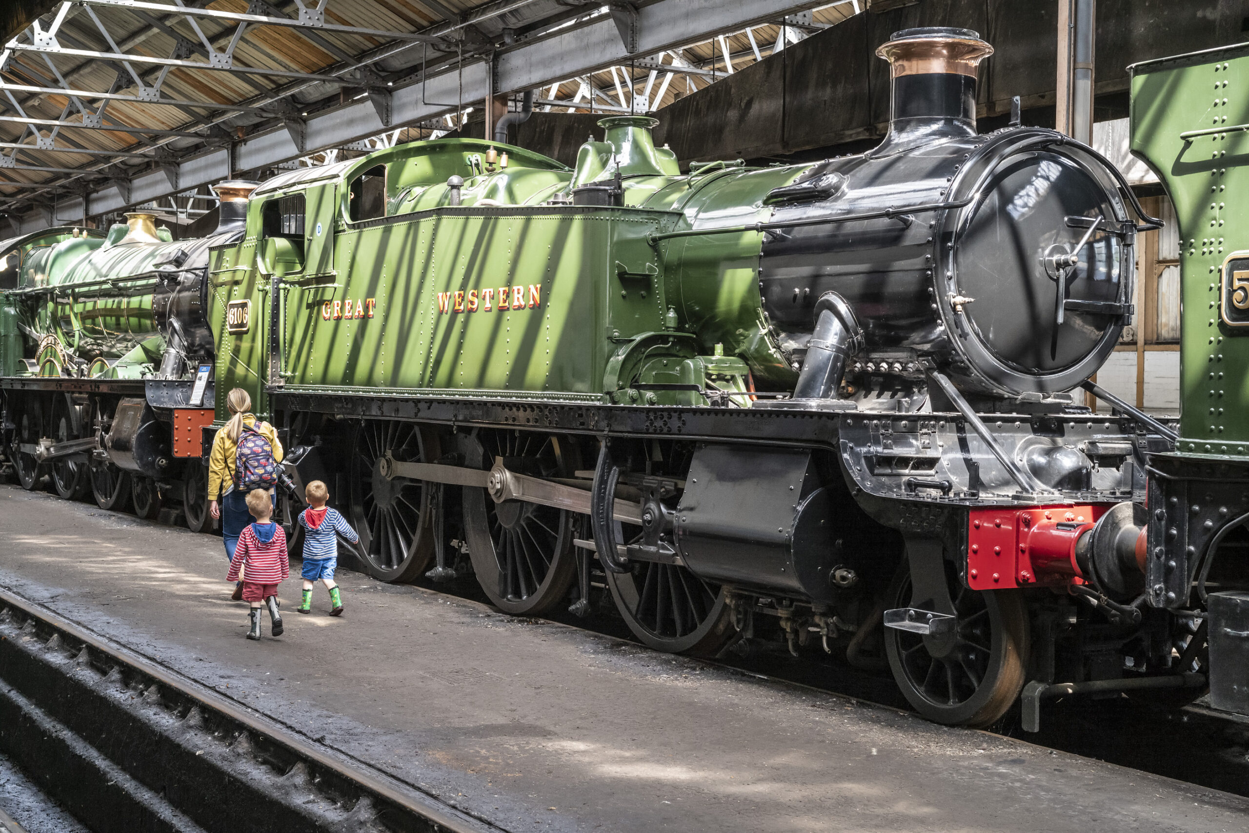 Two small children in striped tops and wellington boots follow a woman with a backpack along a platform beside a gleaming green Great Western steam locomotive, number 6106, inside an engine shed.