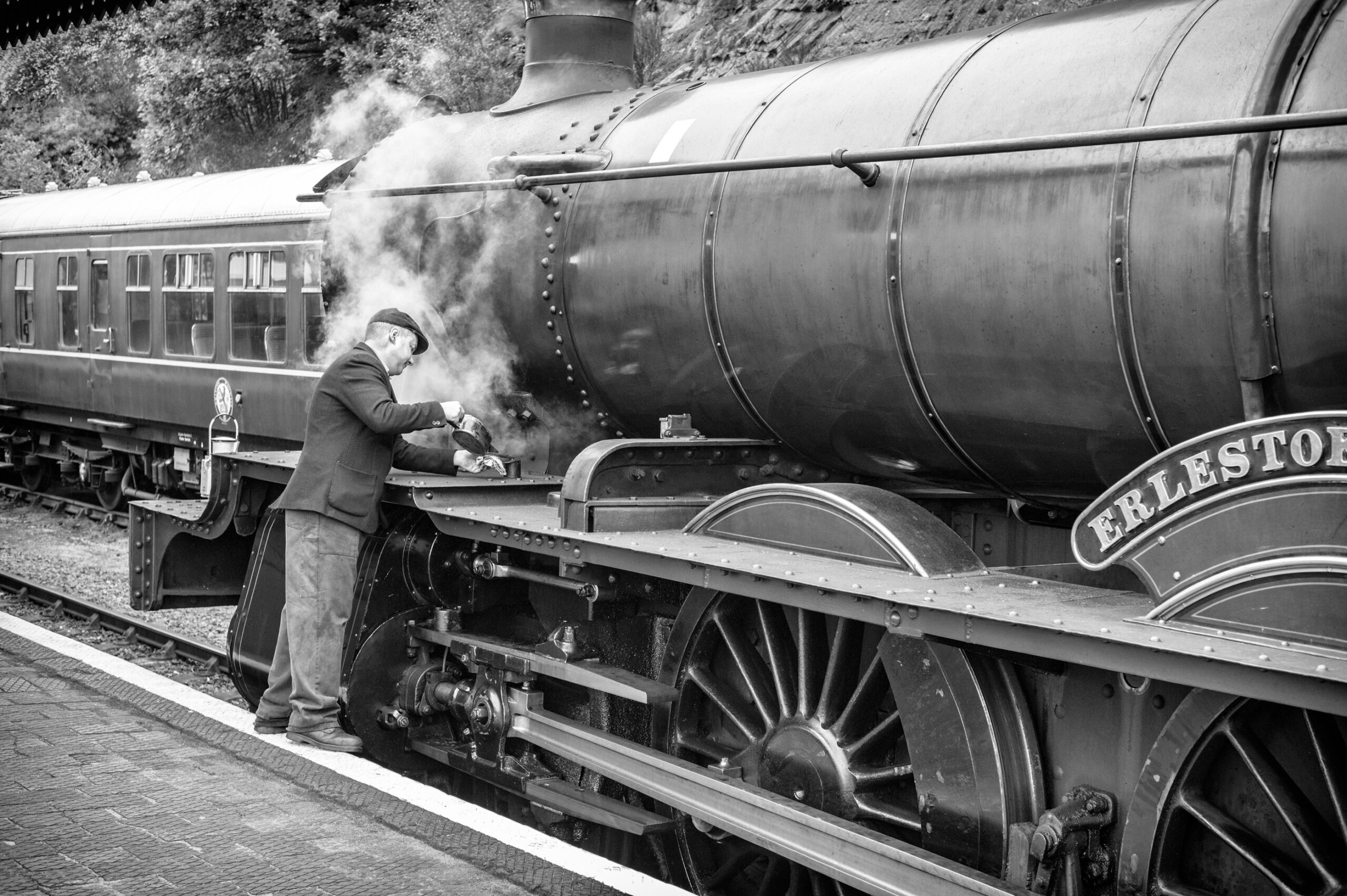Black-and-white photograph of a railwayman in a flat cap oiling a Great Western steam locomotive named Erlestoke Manor as steam drifts around him at a station platform.