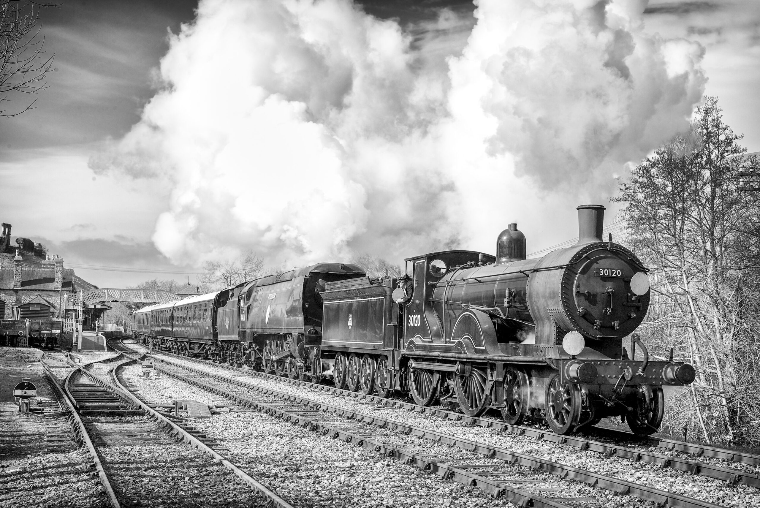 Black-and-white photograph of locomotive 30120 and a
second engine double-heading a train past a country
station, with plumes of steam rising against a cloudy sk