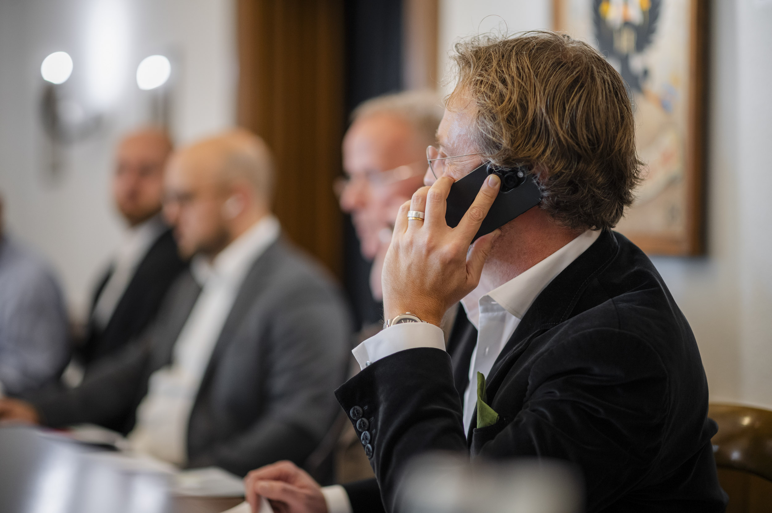 A man in a dark velvet jacket holds a mobile phone to his ear, with other people seated in the background out of focus.