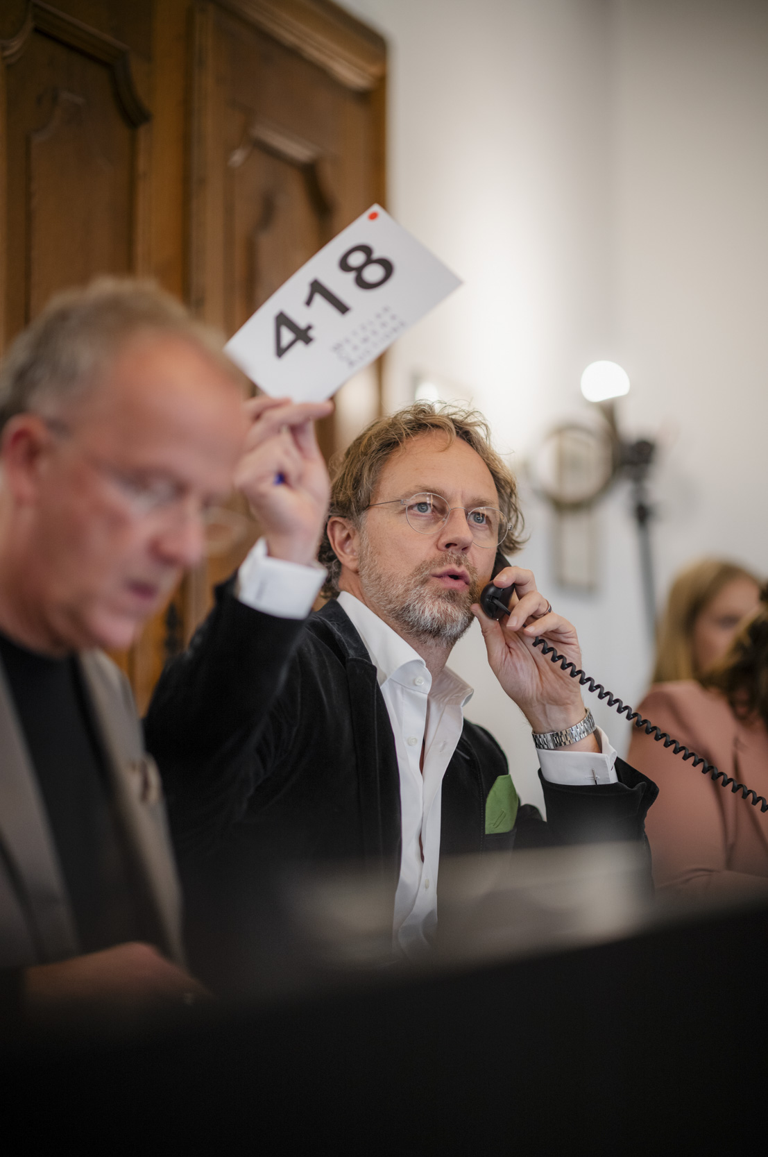 A man with glasses speaks on a corded telephone while holding up bidding paddle number 418.