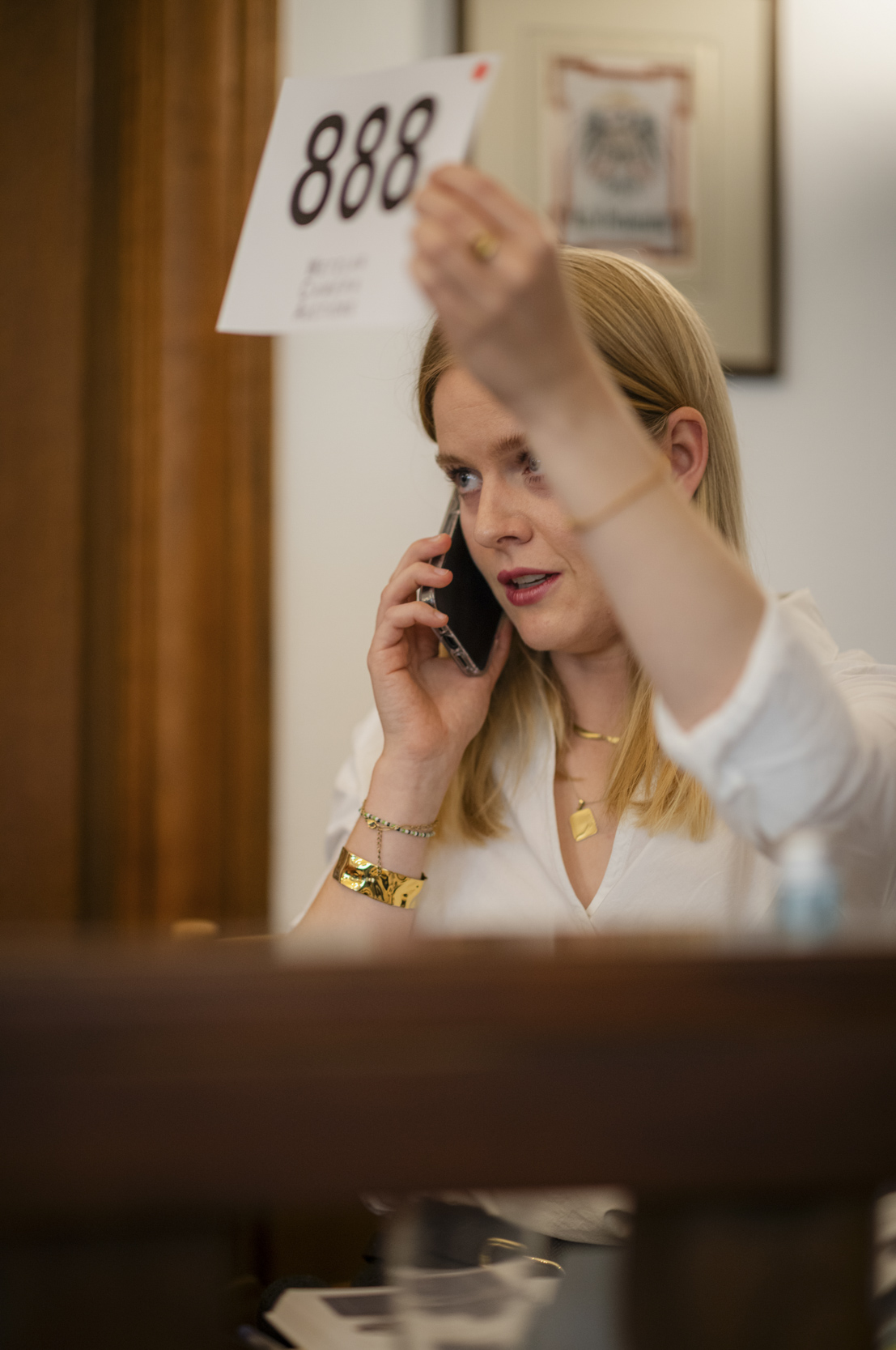A woman with long fair hair speaks on a mobile phone while raising bidding paddle number 888.