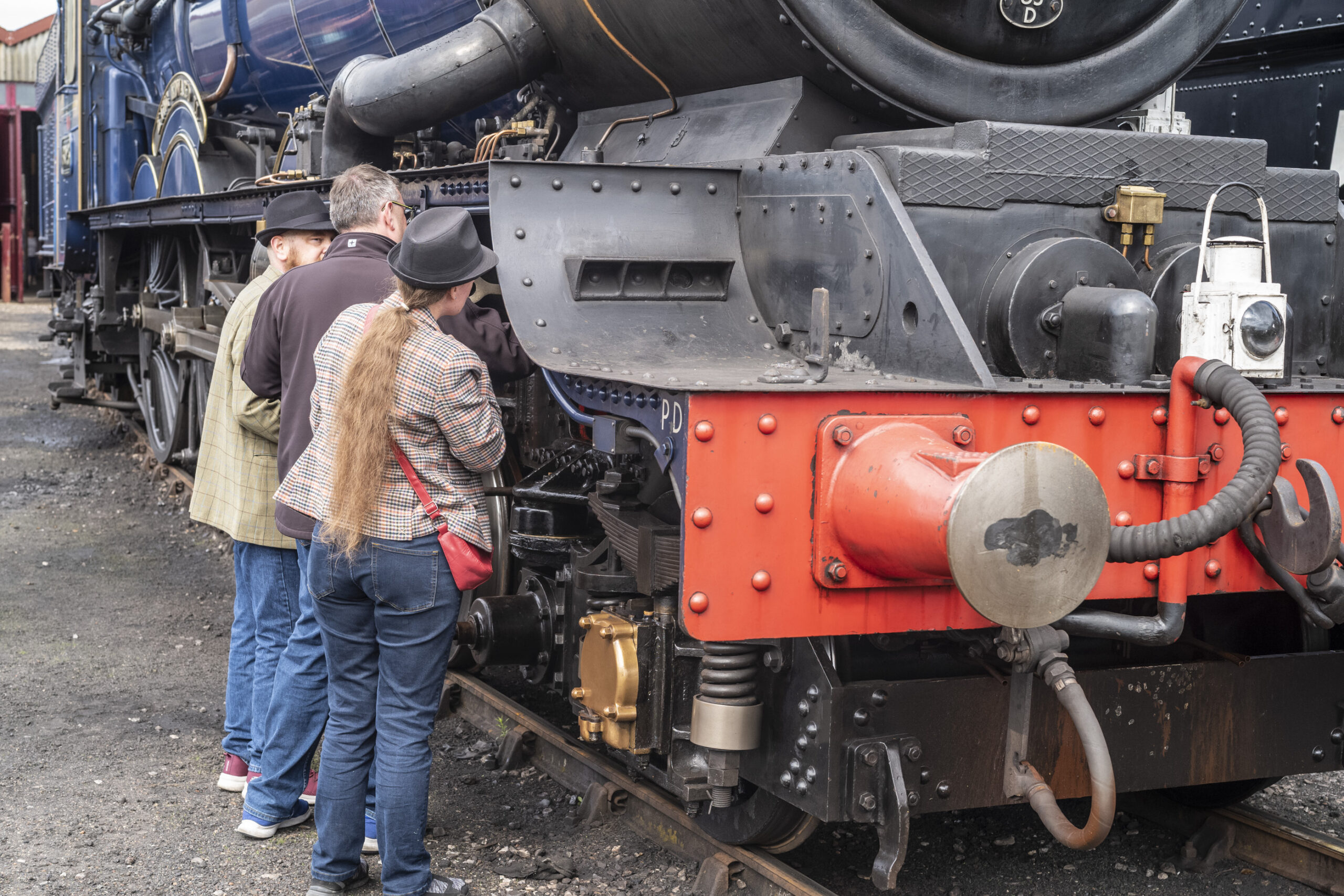 Three visitors, two wearing trilby hats, peer into the inner workings of a steam locomotive beside its bright red buffer beam, with a blue express engine behind.