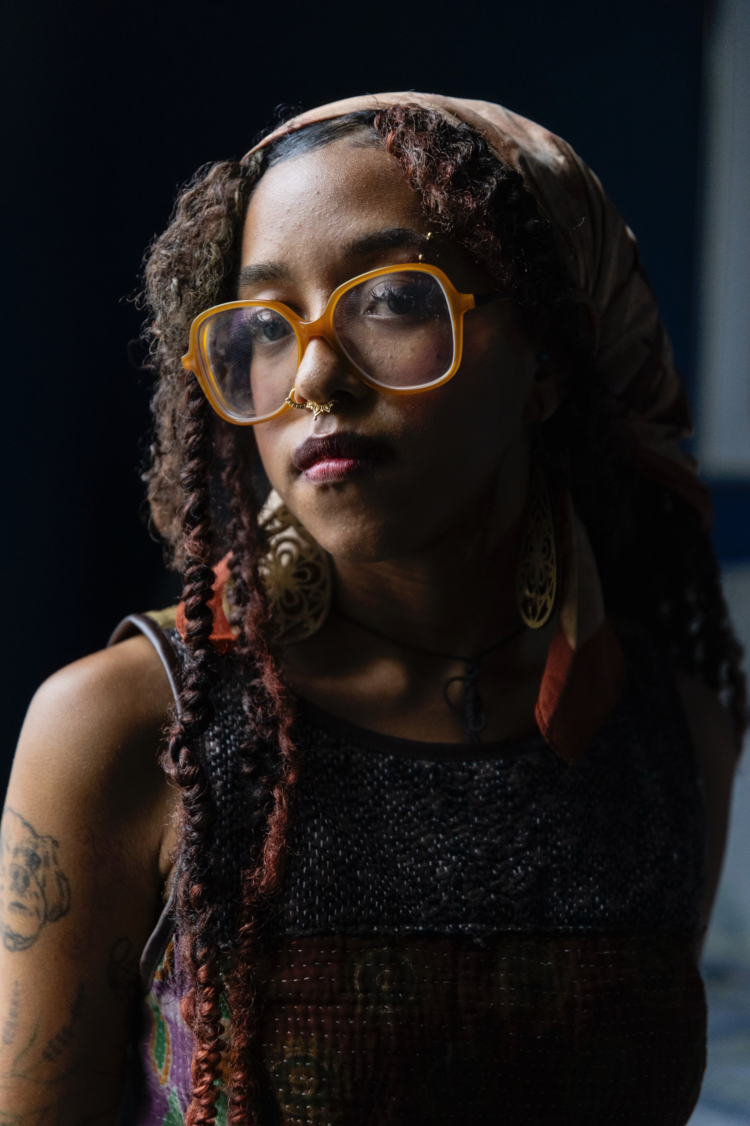 Portrait of Laila AnnMarie Stevens in amber-framed glasses, a patterned headscarf and long twisted braids, lit from one side against a dark background.
