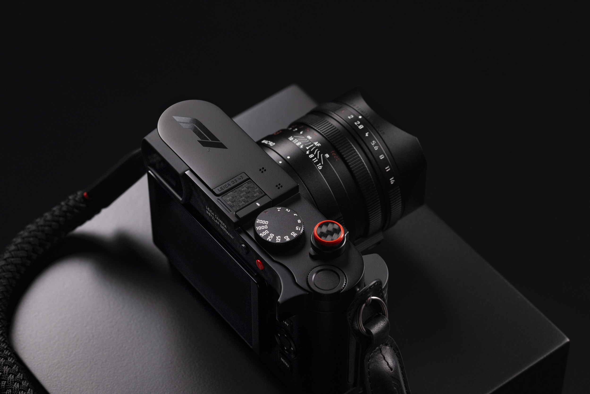 The camera seen from above and behind, showing the F1 logo on the top plate, a carbon fibre hot-shoe cover and the red-rimmed carbon soft-release button, with the rope strap curling in the foreground.