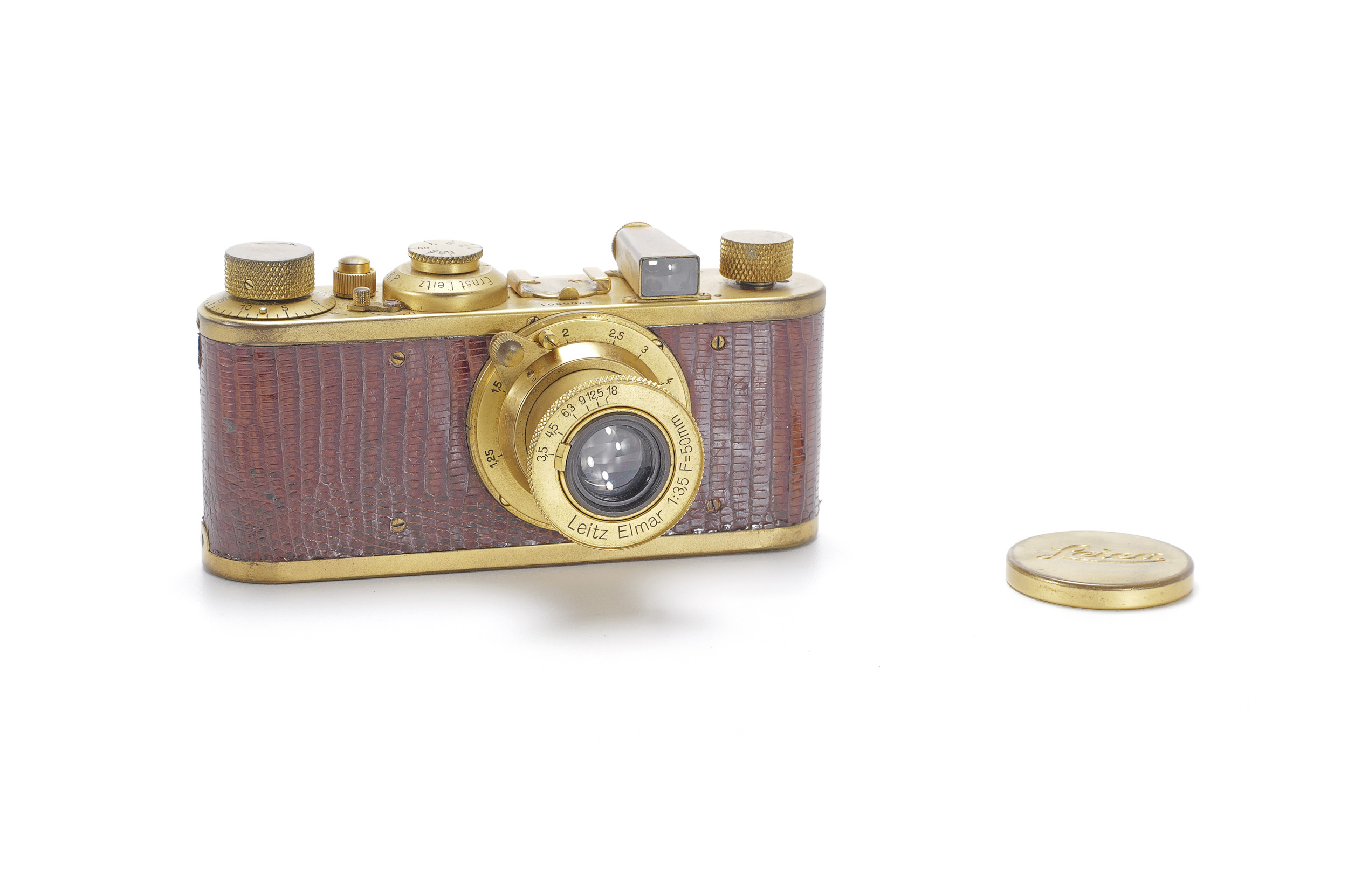 A gold-plated Leica camera covered in brown reptile-skin leather, with a gold Elmar lens and a gold Leica lens cap beside it.