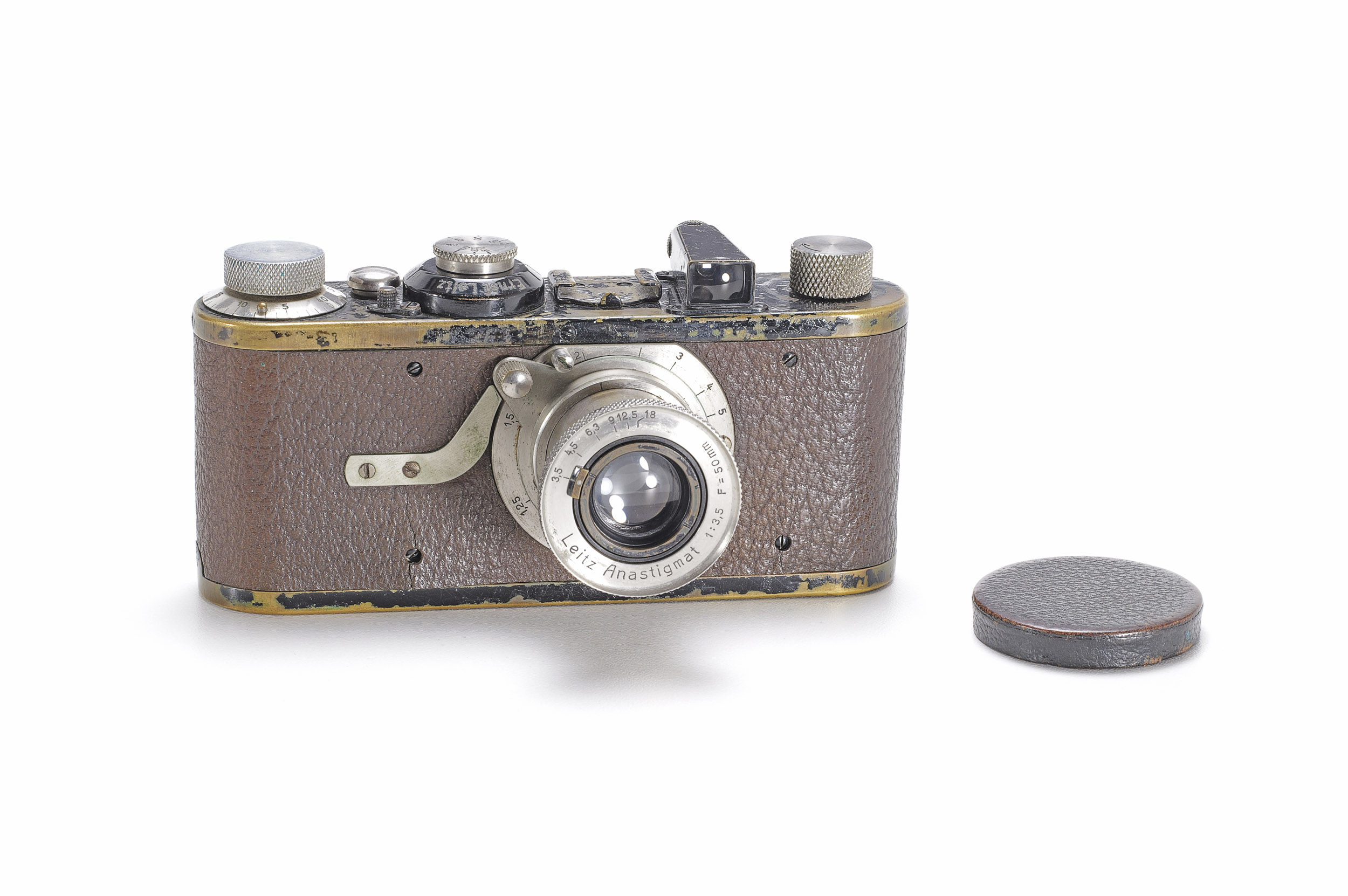 An early Leica camera with brown leather covering, worn black paint showing brass edges, a nickel Anastigmat lens and a leather lens cap.