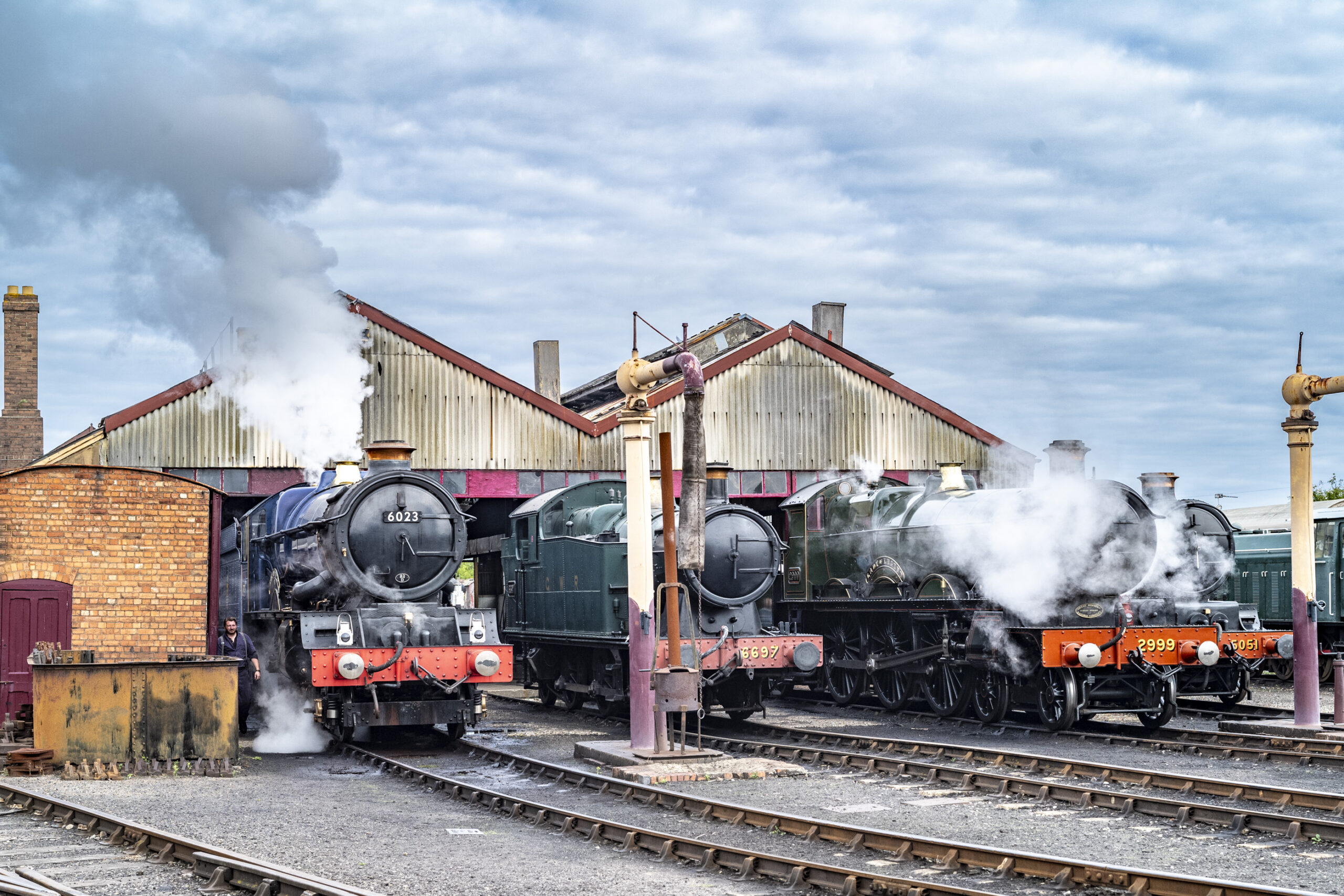 Four steam locomotives, including 6023 and 2999, simmer in clouds of steam outside a corrugated-roofed engine shed beside water cranes under a cloudy sky.