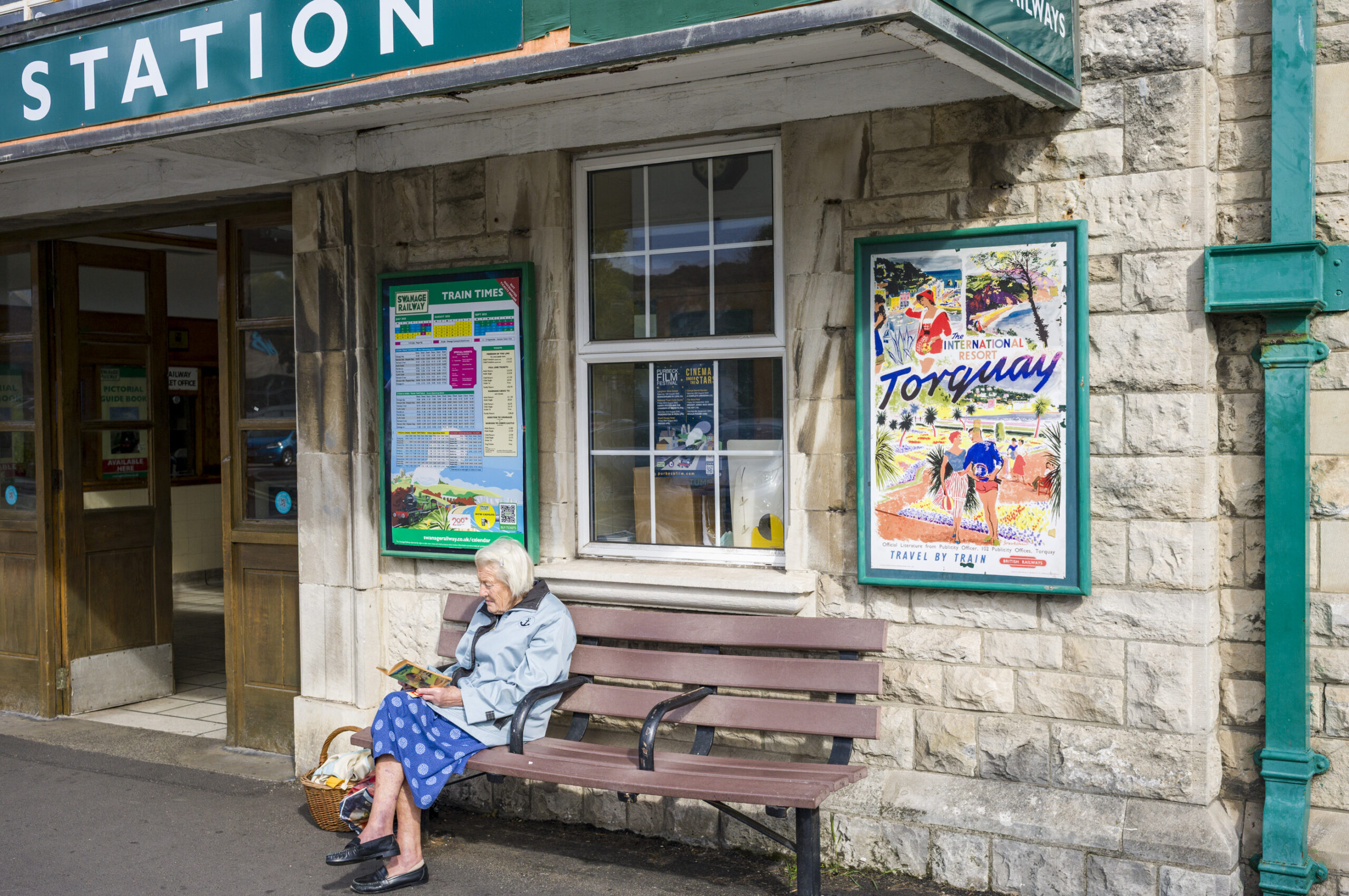 An elderly woman reads on a bench outside a station building, beneath a vintage British Railways poster advertising Torquay and a Swanage Railway timetable.