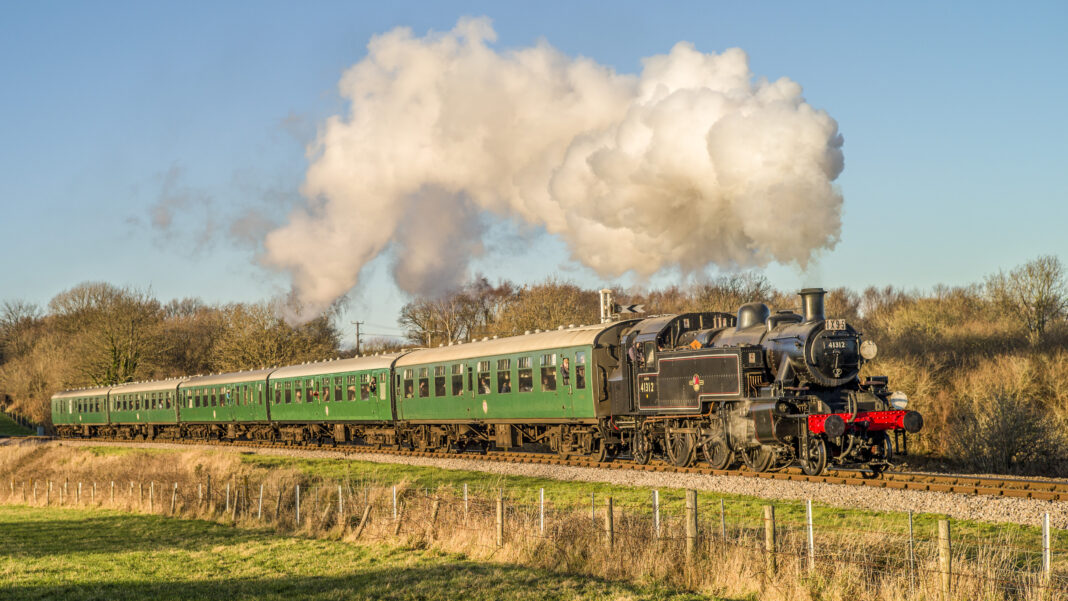 Black tank locomotive 41312 hauls a rake of green carriages through sunlit winter countryside, trailing a huge white plume of steam against a blue sky.