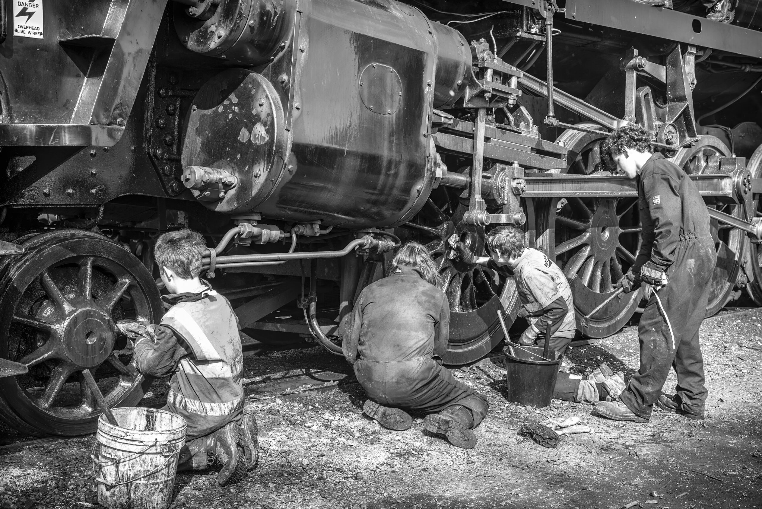 Black-and-white photograph of four young volunteers in grimy overalls cleaning the wheels and motion of a large steam locomotive with buckets and rags.