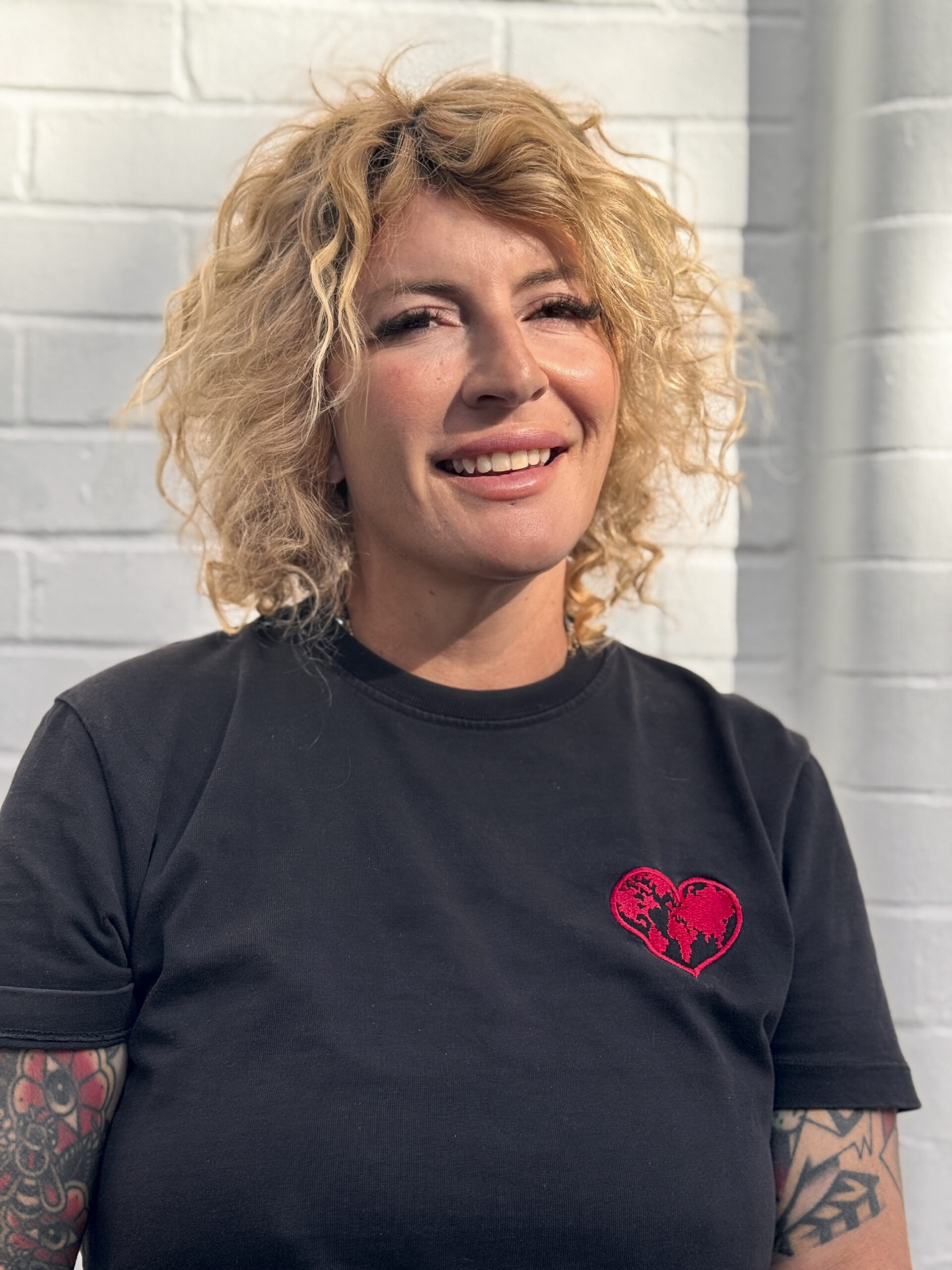Portrait of Valentina Sinis, smiling, with curly blonde hair and tattooed arms, in a black T-shirt with an embroidered red heart, against a white-painted brick wall.